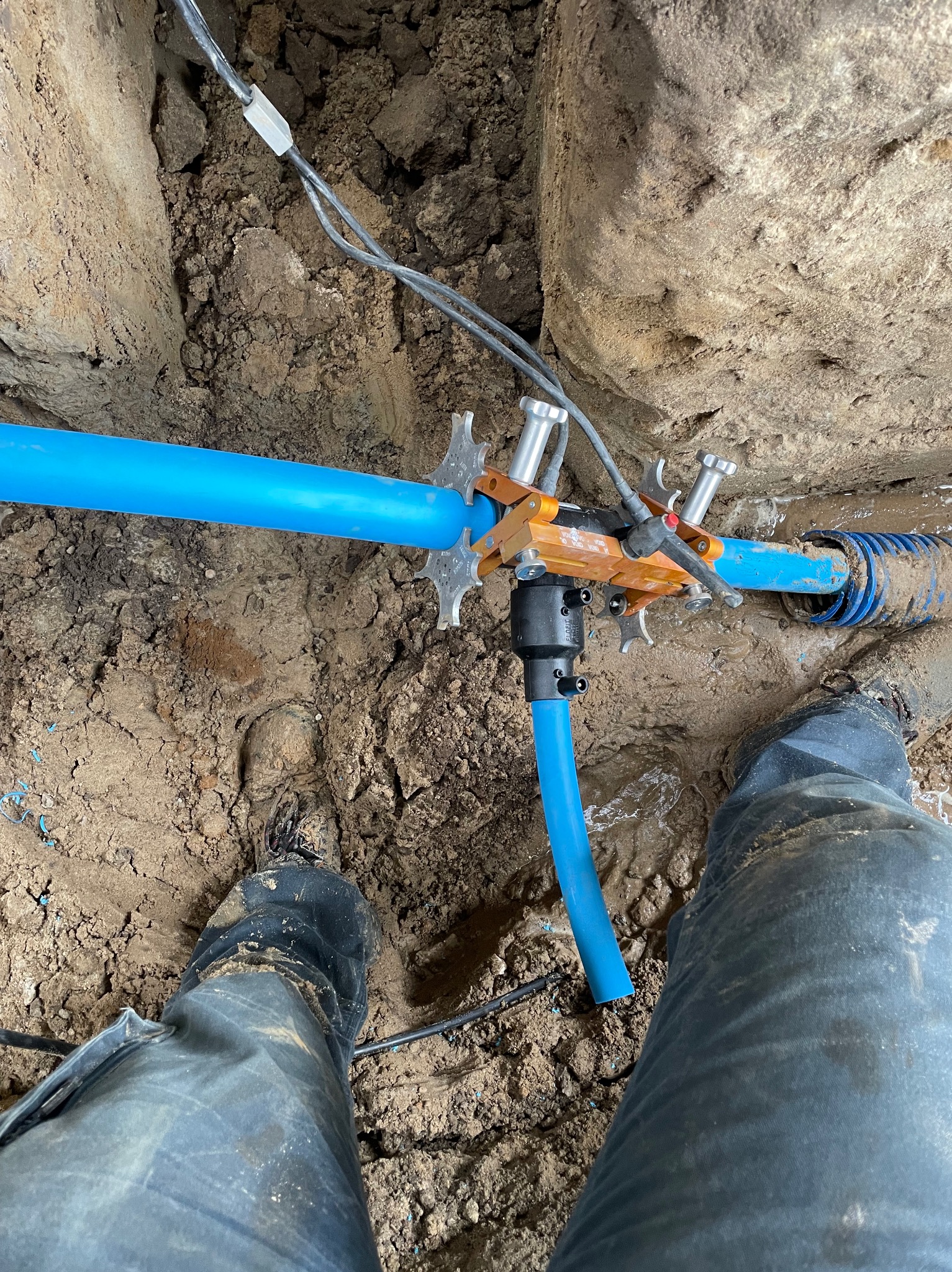 Niebieskie rury wodociągowe łączone specjalistycznym zaciskiem w wykopie ziemnym, widok z góry na stopy w roboczych butach. Prace instalacyjne w błotnistym terenie.
