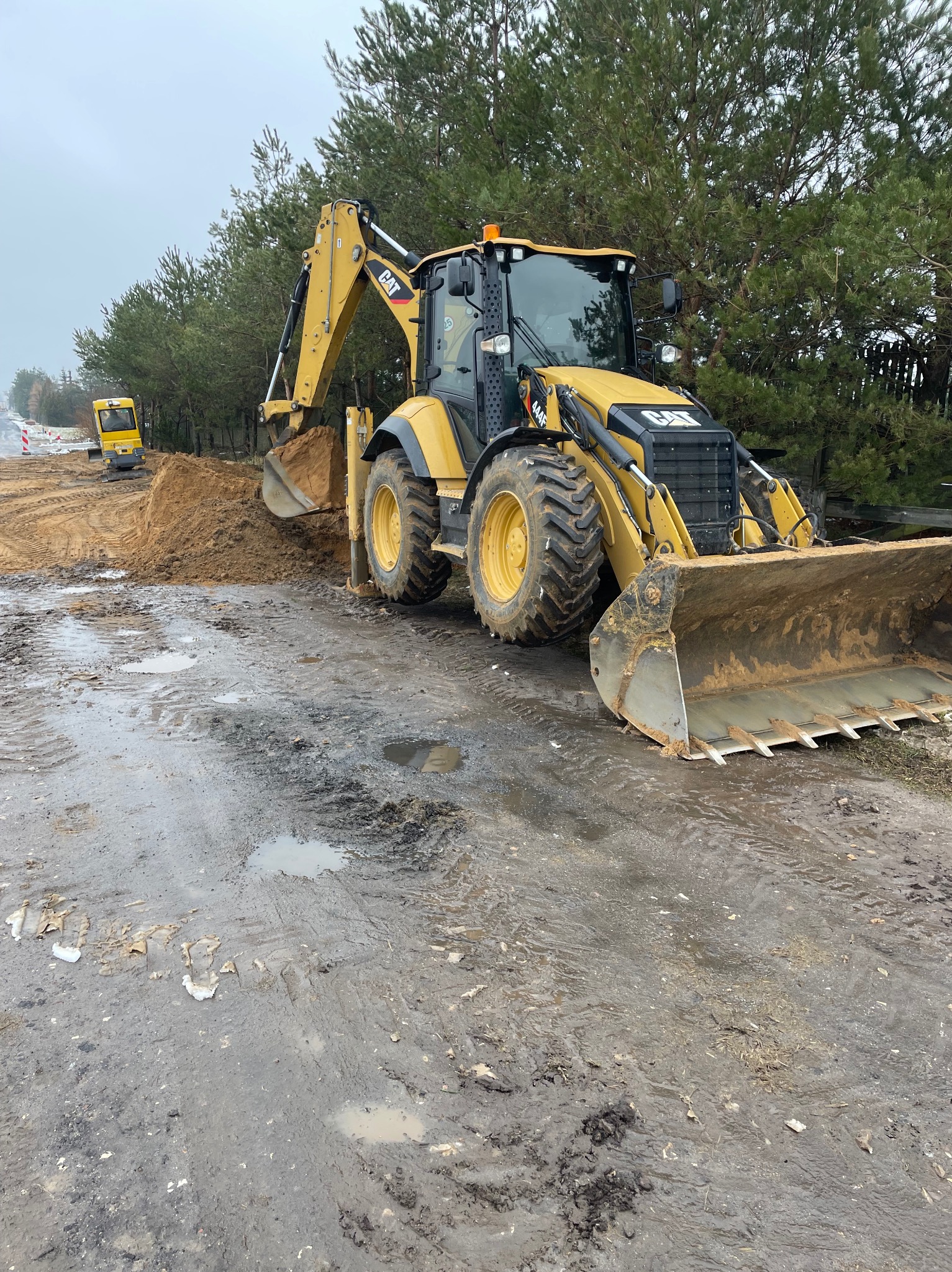 Żółta koparko-ładowarka CAT na błotnistym terenie z wykopaną ziemią, w tle las i mniejsza maszyna budowlana. Praca w toku, pochmurny dzień.