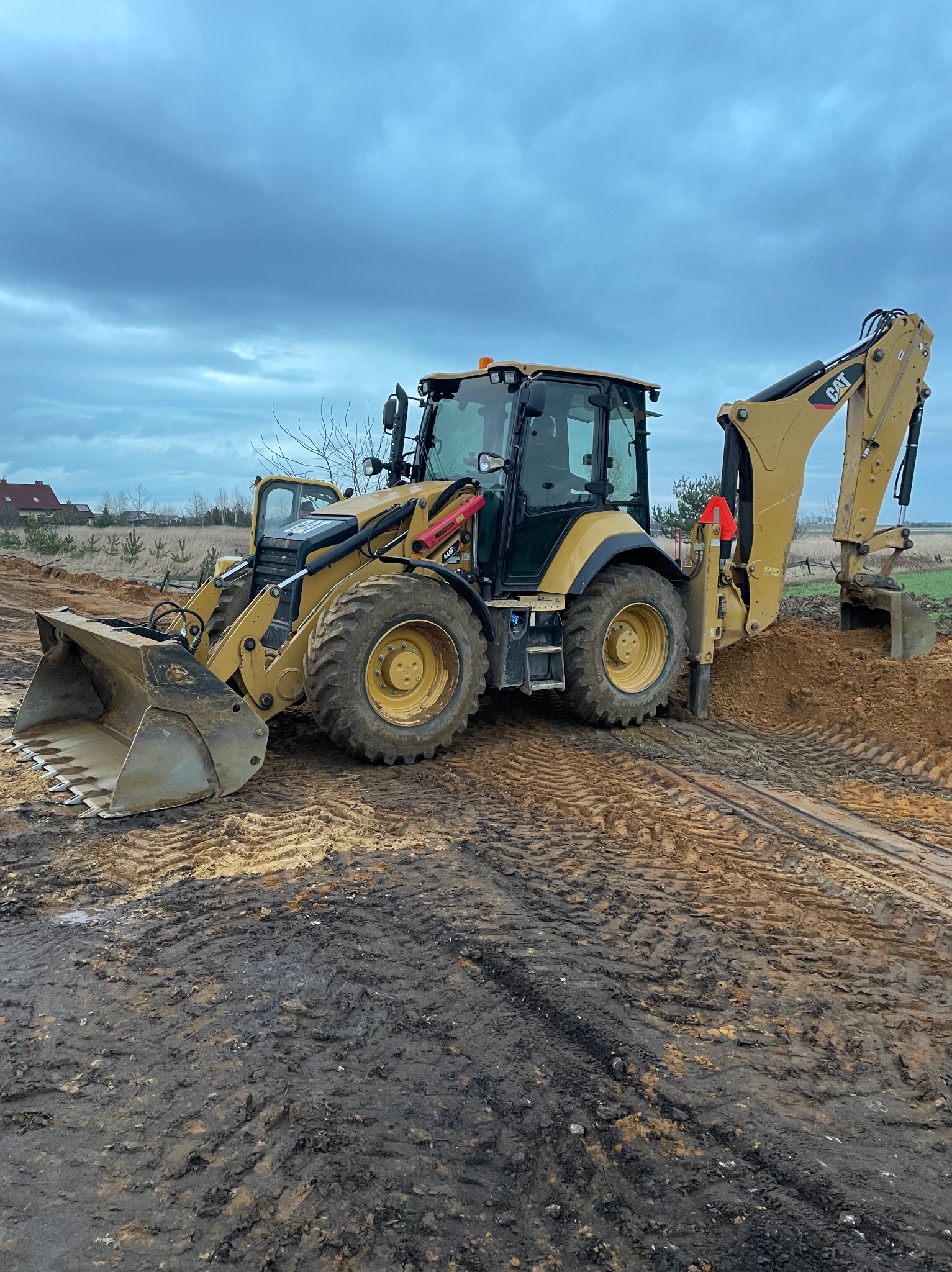 Żółta koparko-ładowarka CAT na placu budowy, z łyżką zagłębiającą się w ziemię. Widoczne ślady opon w błocie i pochmurne niebo w tle. Praca w toku.