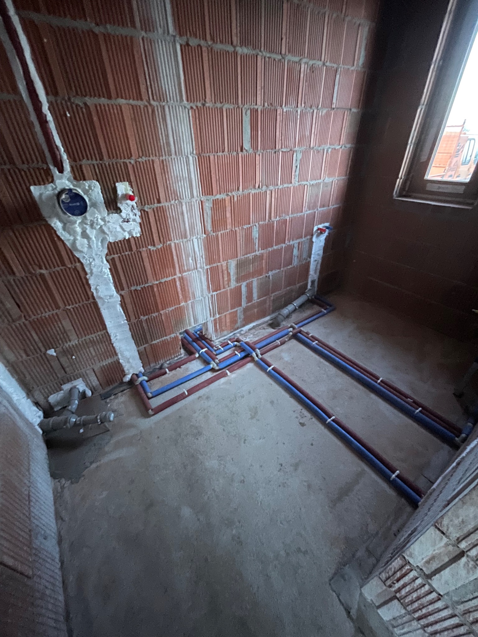 Wnętrze w trakcie prac hydraulicznych: czerwona cegła, rury wodne (niebieskie i czerwone) prowadzone po podłodze, instalacja zabezpieczona pianką montażową przy ścianie.