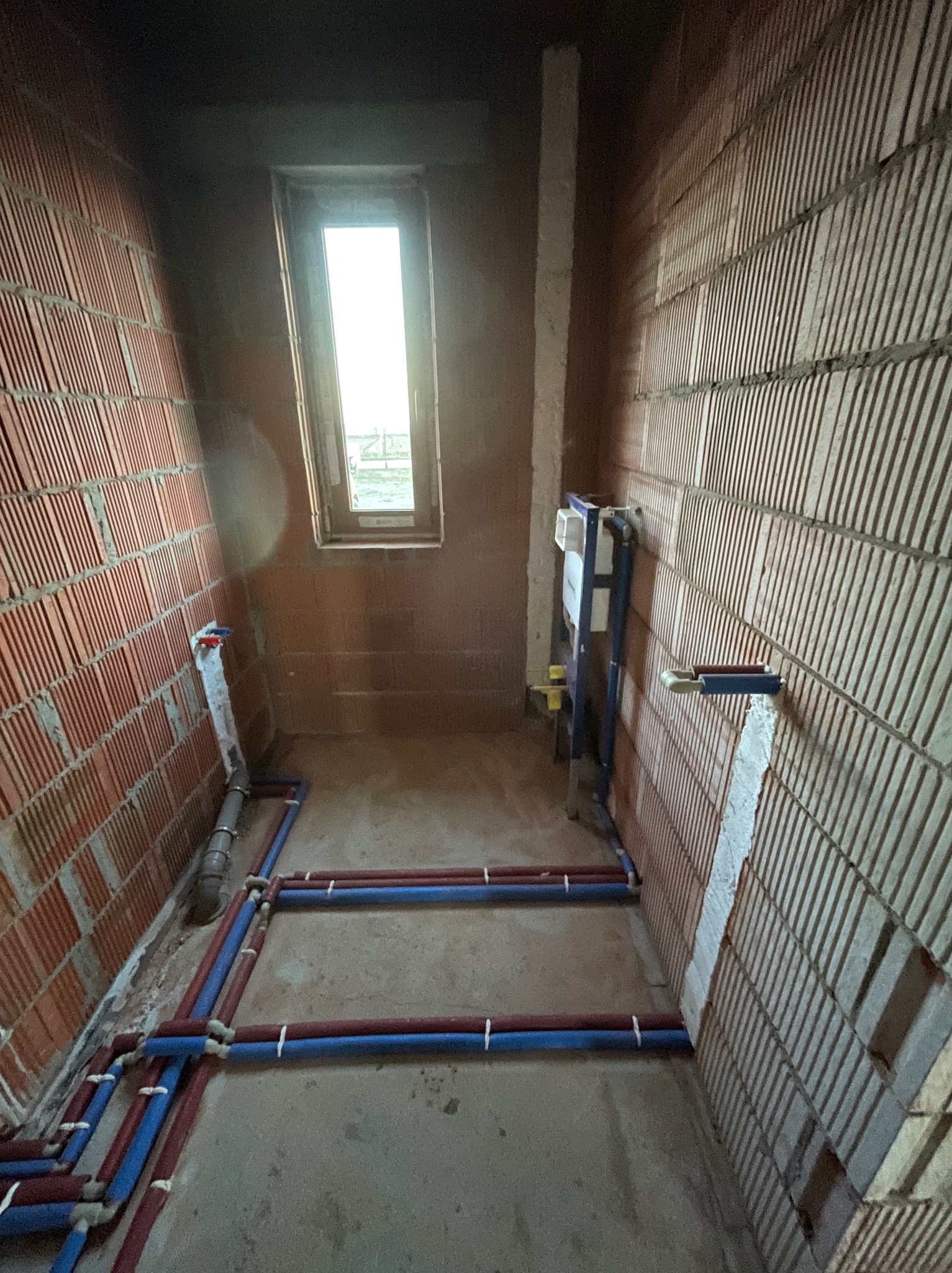 Surowy stan łazienki z widoczną instalacją wodną z rur w kolorze niebieskim i czerwonym, zamontowaną na ścianie z cegły dziurawki. Widoczne okno i fragment instalacji podtynkowej.