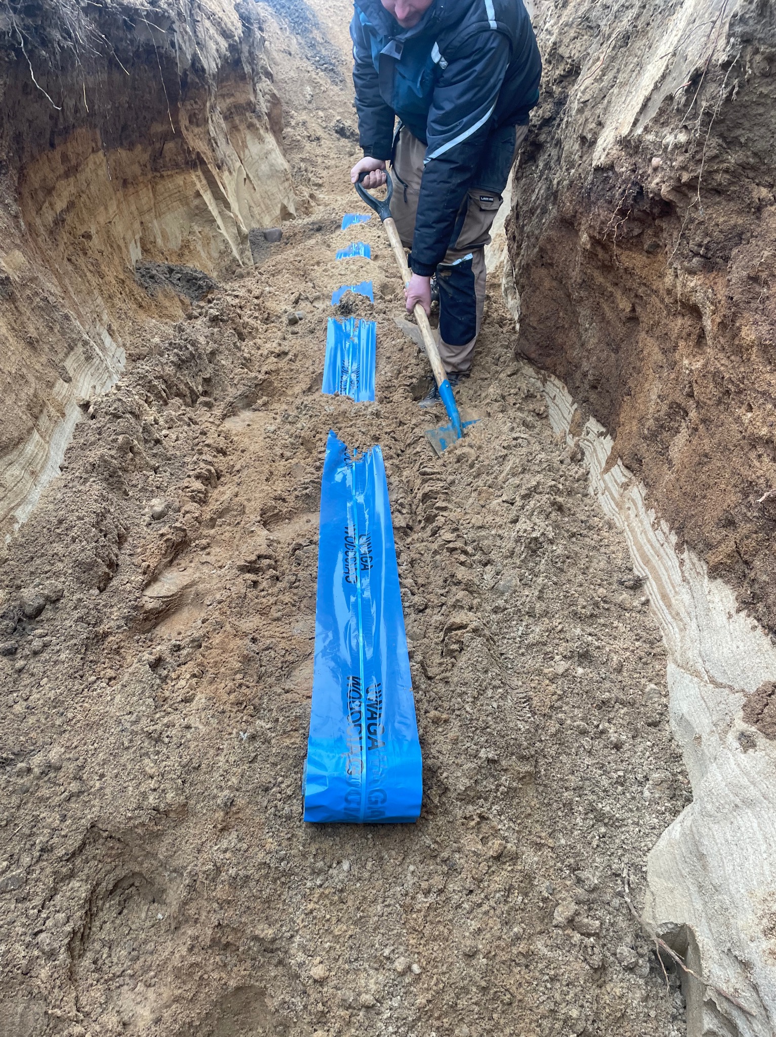 Wykop w ziemi z niebieską folią ostrzegawczą ułożoną wzdłuż wykopu. Pracownik kopie dół łopatą. Widoczne warstwy ziemi w przekroju.