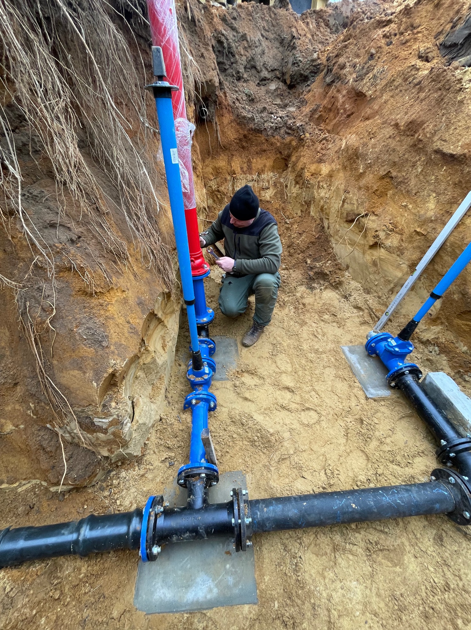 Instalacja wodociągowa w wykopie: hydraulik łączy rury z niebieskimi kołnierzami, czerwona rura w folii. Widoczne narzędzia i poziomica.