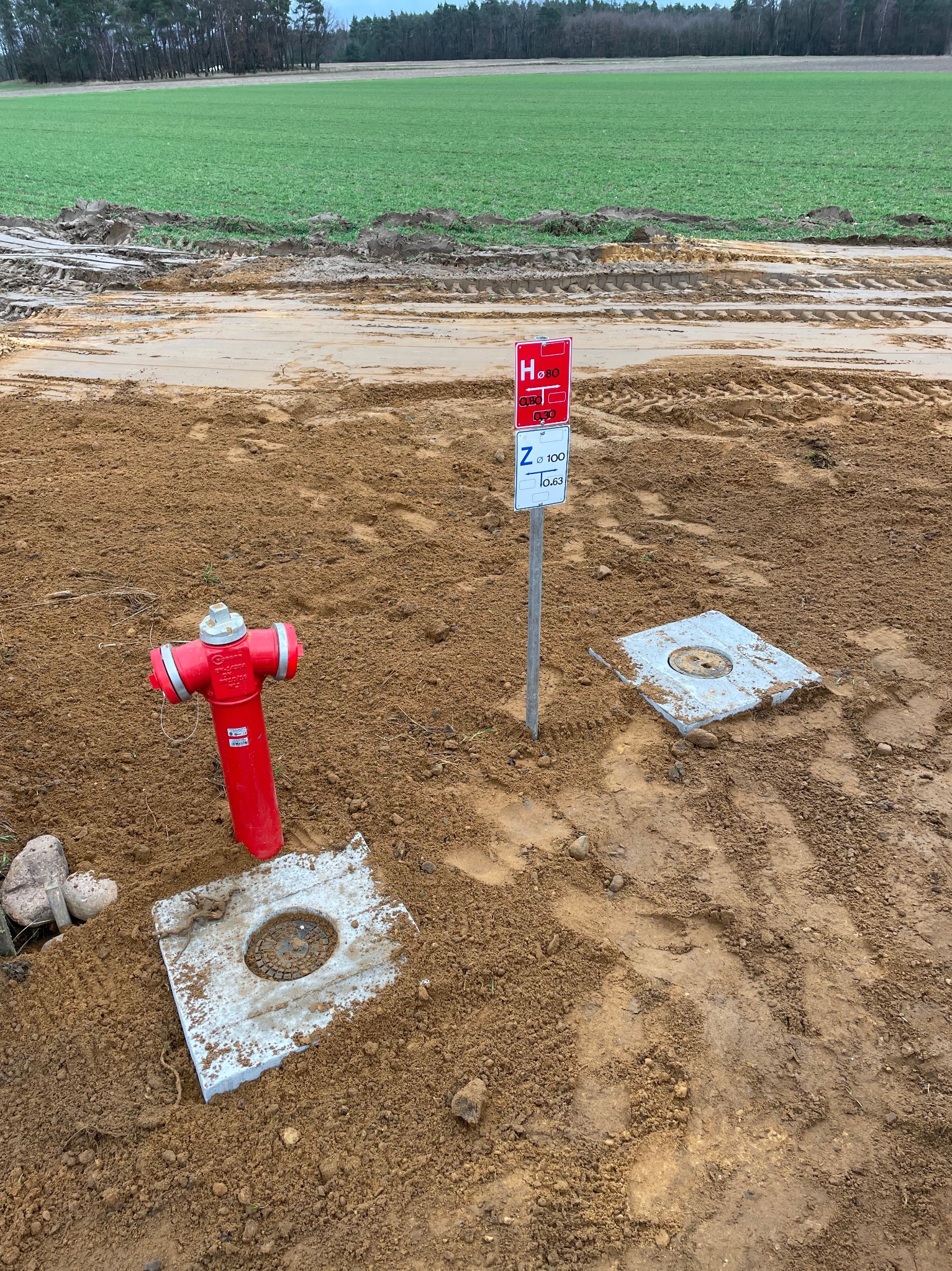 Czerwony hydrant nadziemny i betonowe włazy na ziemi, z tabliczką informacyjną w tle, na tle pola i lasu. Widoczne ślady opon.