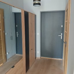 ProCraftHouse - Nowoczesny przedpokój z drewnianą szafą z lustrem i szarą ścianą. Widoczne dwoje drzwi: wewnętrzne drewniane i zewnętrzne szare. Minimalistyczna lampa sufitowa.