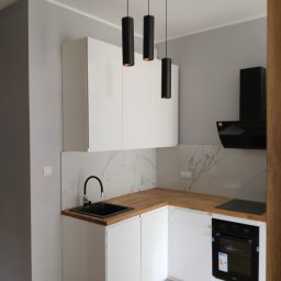 ProCraftHouse - Biała, nowoczesna kuchnia z drewnianym blatem i marmurowym backsplashiem. Czarne, cylindryczne lampy wiszące nad zlewem. Minimalistyczny design.