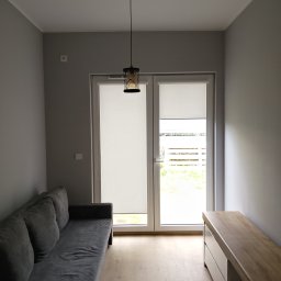 ProCraftHouse - Jasne wnętrze po remoncie: szara sofa, drewniana podłoga, białe drzwi balkonowe z roletami i lampa wisząca. Minimalistyczny design.