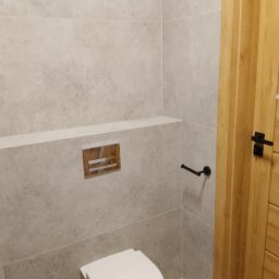 ProCraftHouse - Jasna łazienka z szarymi płytkami imitującymi kamień, drewnianymi drzwiami z czarną klamką i chromowanym przyciskiem spłuczki. Minimalistyczny design.