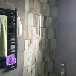 ProCraftHouse - Ściana w łazience wyłożona pionowymi, błyszczącymi płytkami w odcieniach beżu i szarości. Obok grzejnik drabinkowy z ręcznikami. Widoczny fragment ściany z szarego kamienia.