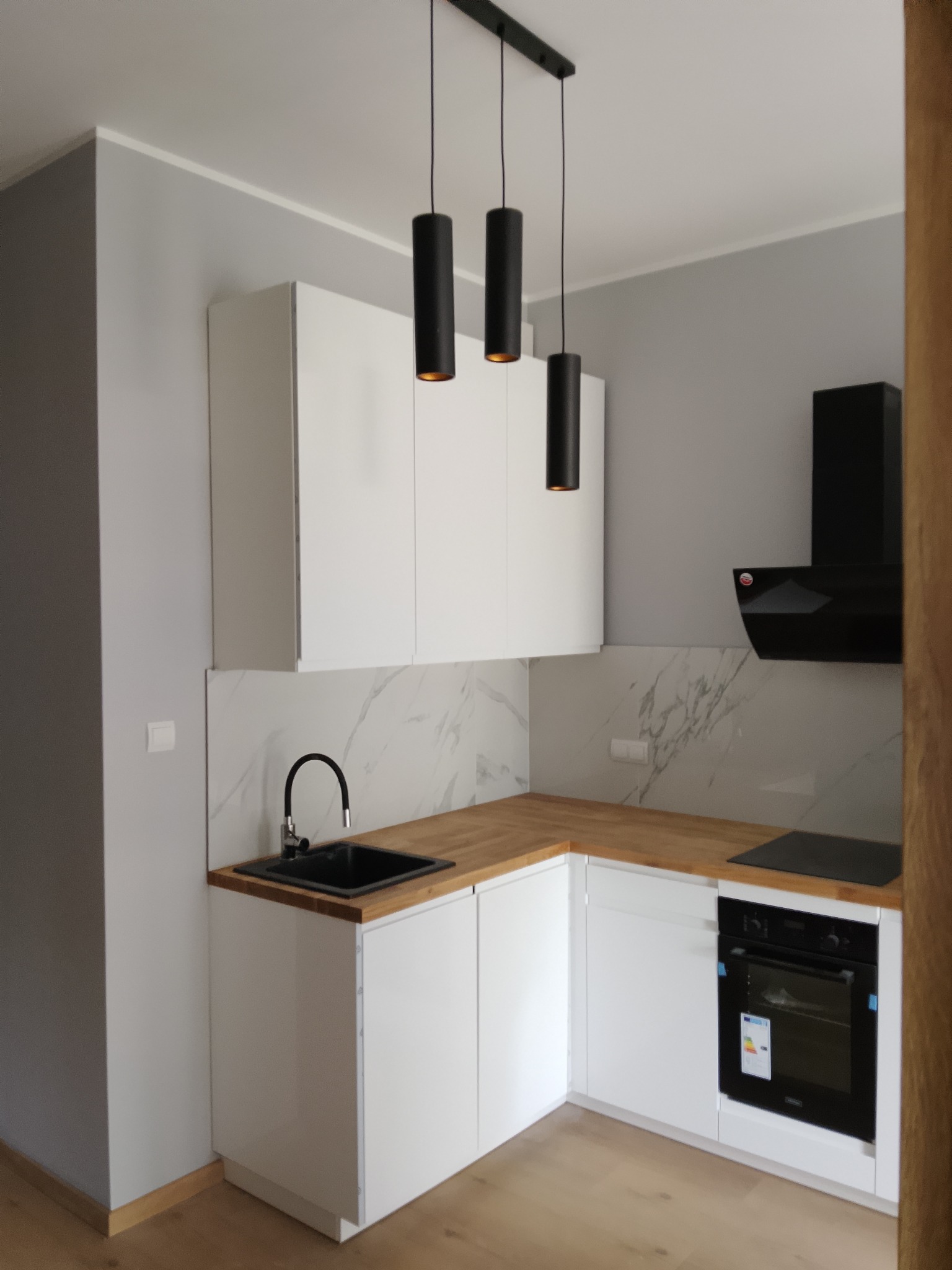 Biała, nowoczesna kuchnia z drewnianym blatem i marmurowym backsplashiem. Czarne, cylindryczne lampy wiszące nad zlewem. Minimalistyczny design.