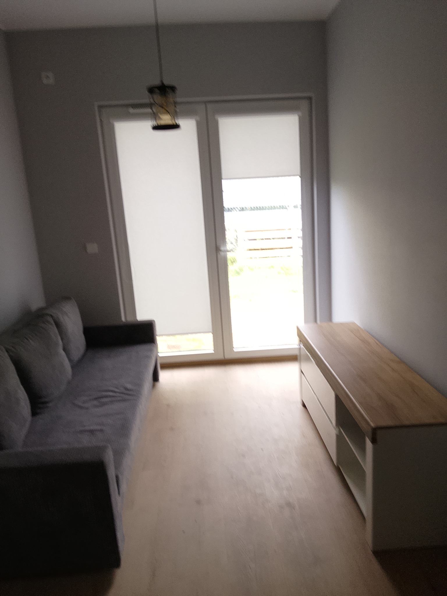 Jasne wnętrze pokoju po remoncie: szara sofa, drewniana podłoga, biała komoda z drewnianym blatem i okno balkonowe z roletami. Minimalistyczny wystrój.