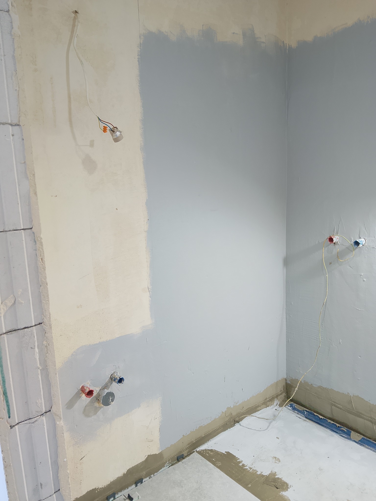 Ściana w trakcie malowania na szaro, widoczne instalacje elektryczne i hydrauliczne. Częściowo pomalowana ściana w rogu pomieszczenia, przygotowania do wykończenia.