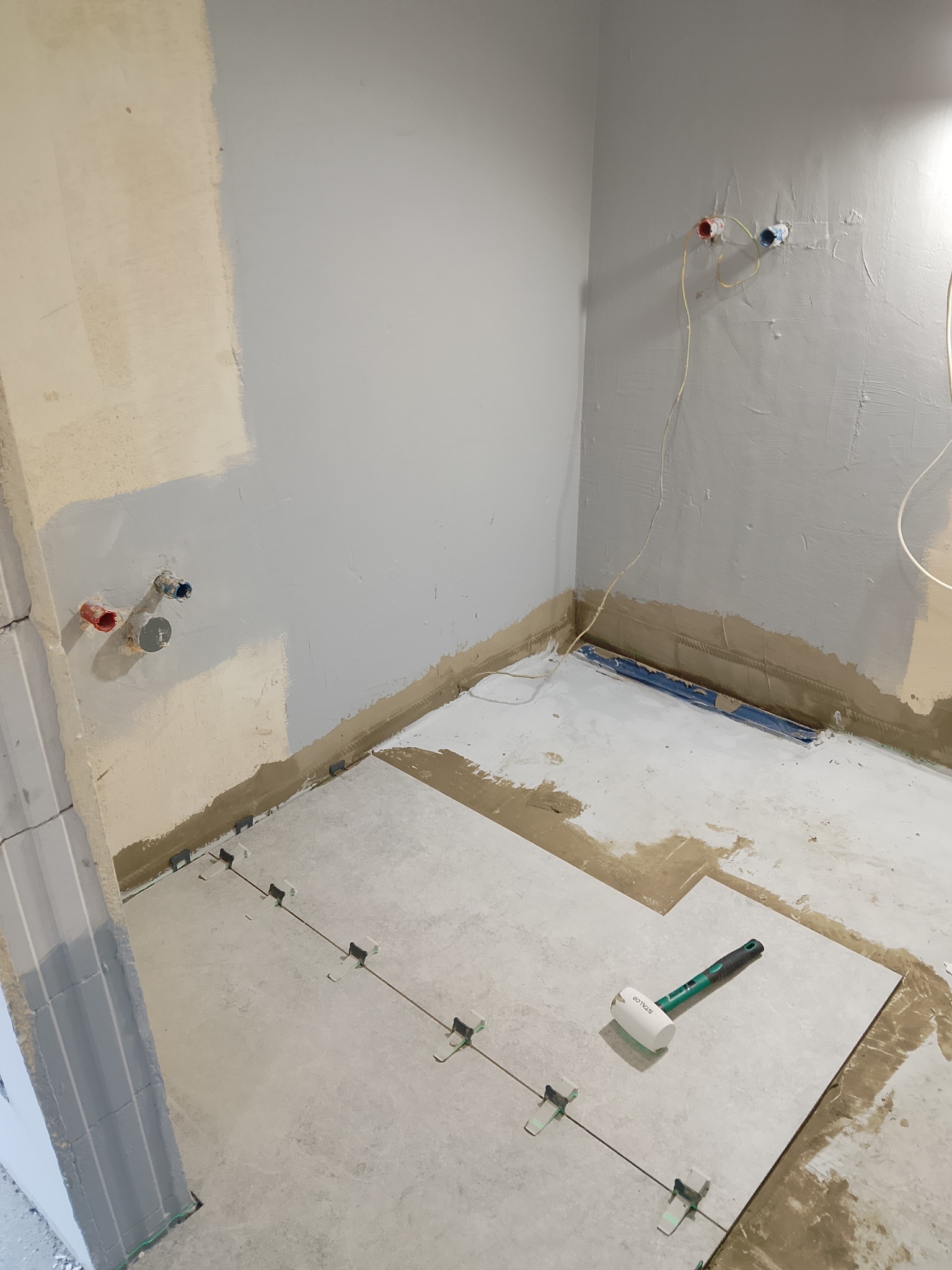 Układanie jasnoszarych płytek podłogowych w łazience, widoczne krzyżyki dystansowe i gumowy młotek. Ściany częściowo przygotowane do dalszych prac.