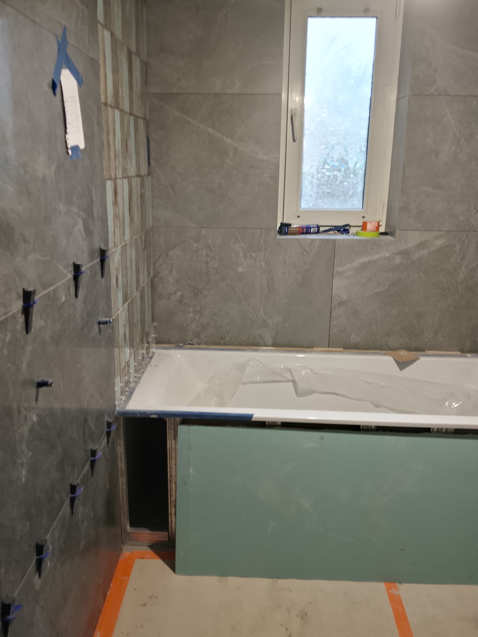 Montaż płytek w łazience z wanną. Widoczne narzędzia glazurnicze i fragment okna. Szare płytki imitujące kamień. Prace w toku, zabezpieczona wanna.