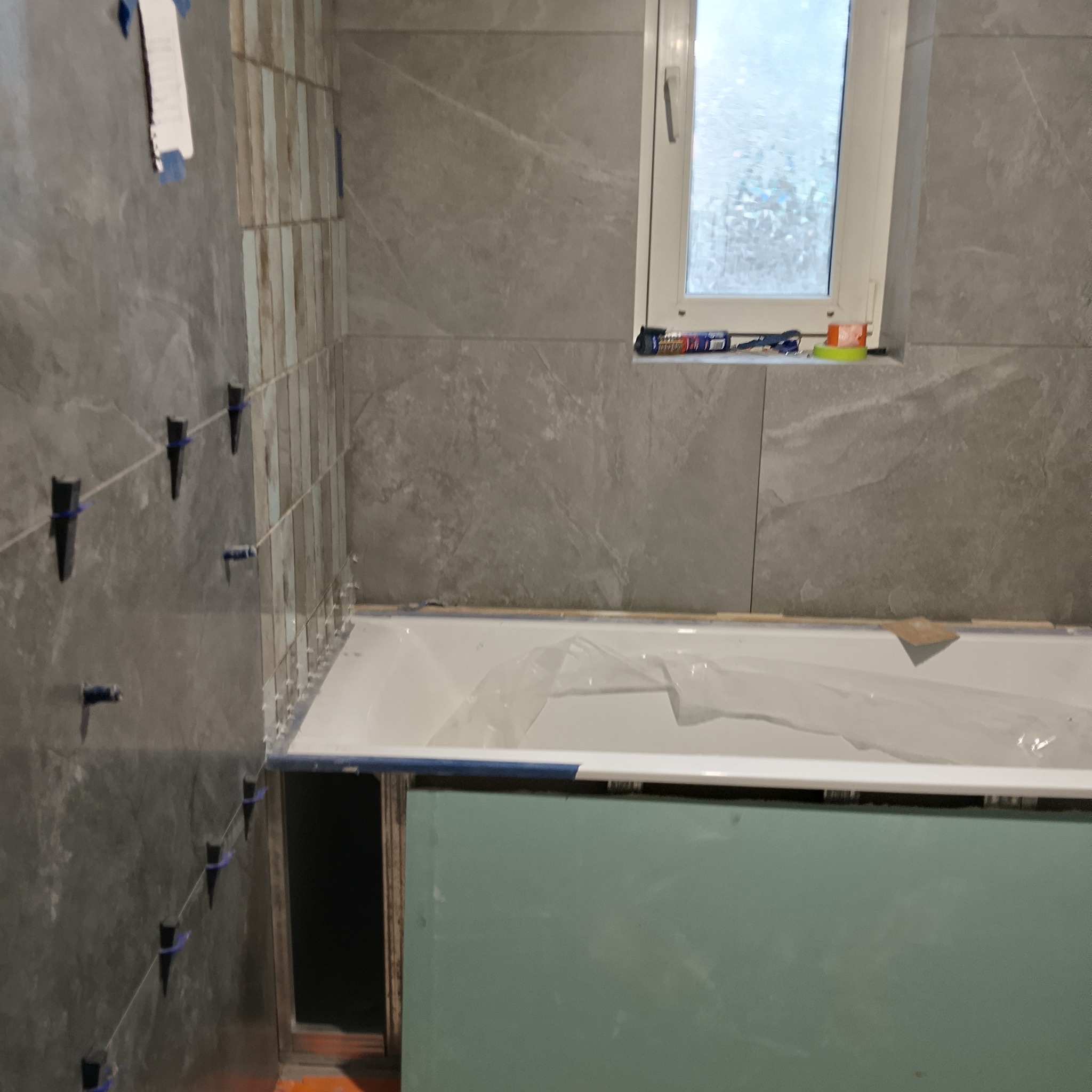 Układanie płytek w łazience w Żyrardowie. Widoczne szare płytki imitujące marmur, krzyżyki dystansowe i przygotowana obudowa wanny z płyty GK.