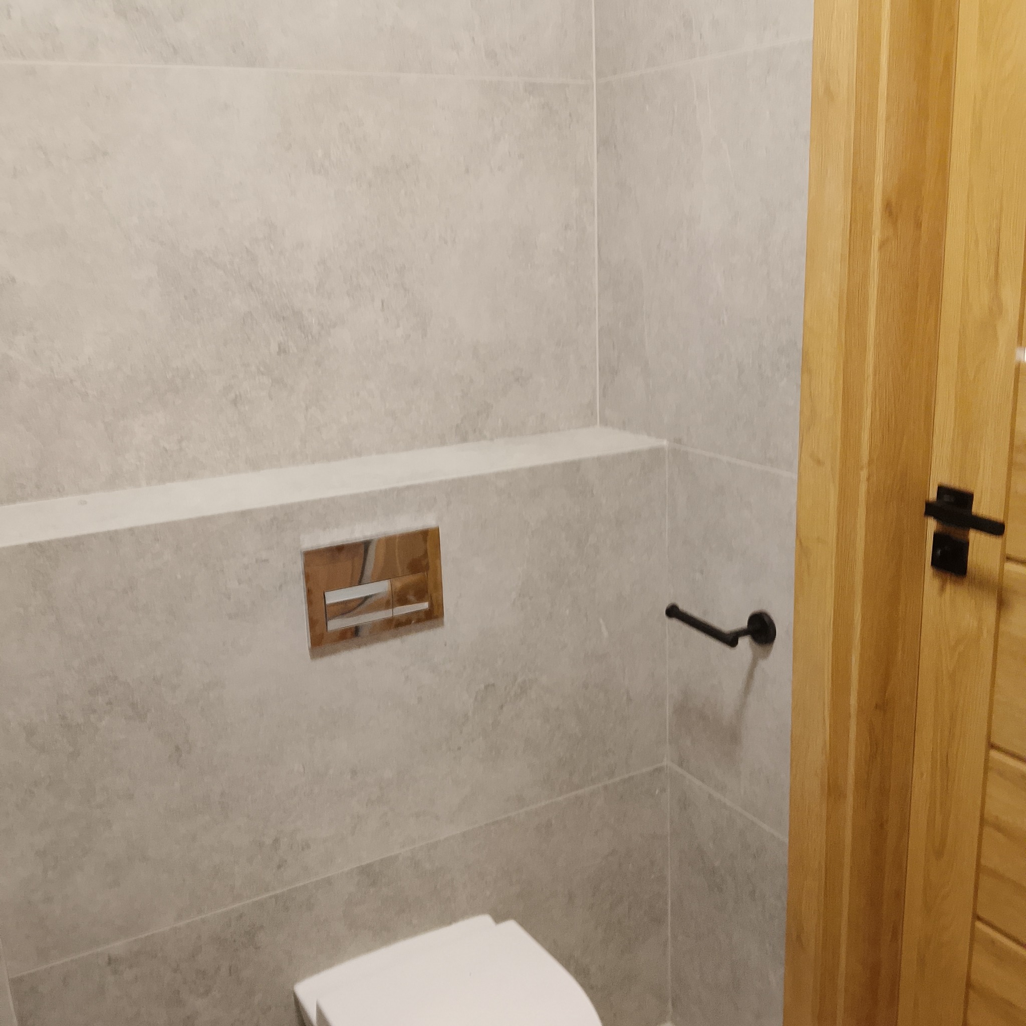 Jasna łazienka z szarymi płytkami imitującymi kamień, drewnianymi drzwiami z czarną klamką i chromowanym przyciskiem spłuczki. Minimalistyczny design.