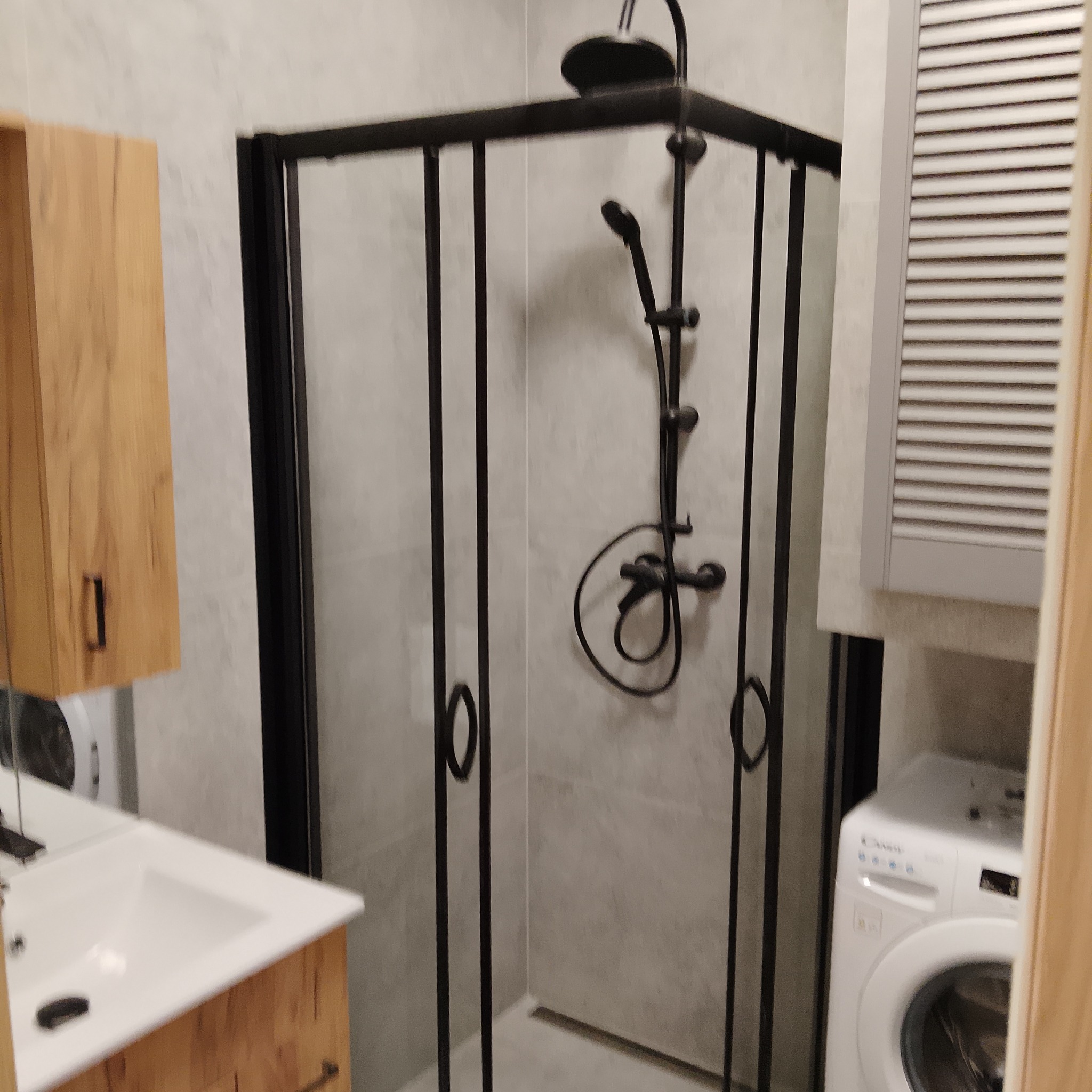 Nowoczesna kabina prysznicowa z czarną armaturą w wykończonej łazience. Widoczna umywalka z szafką z drewnianym frontem i pralka. Minimalistyczny design.