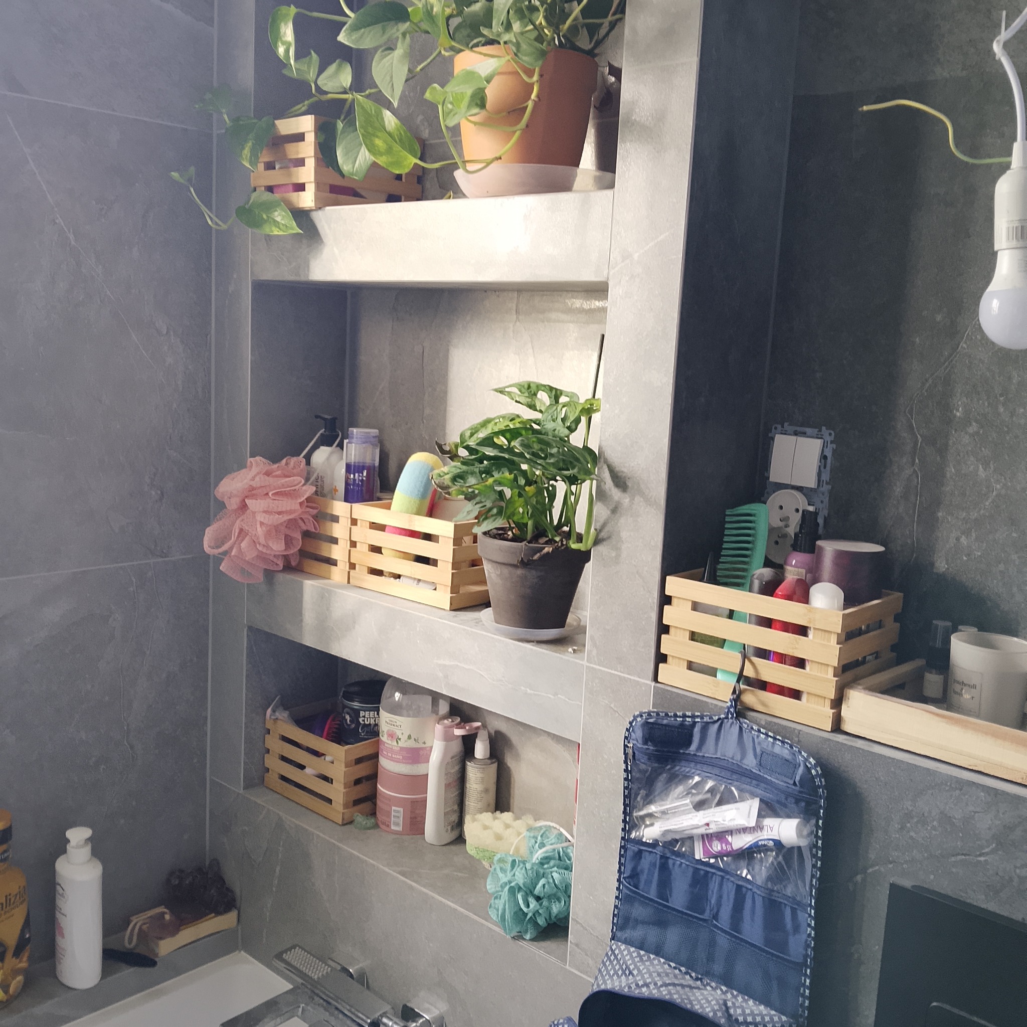 Nowoczesna łazienka z szarymi płytkami i wnękami, w których umieszczono drewniane pojemniki z kosmetykami i roślinami doniczkowymi. Minimalistyczny design, ciepłe oświetlenie.