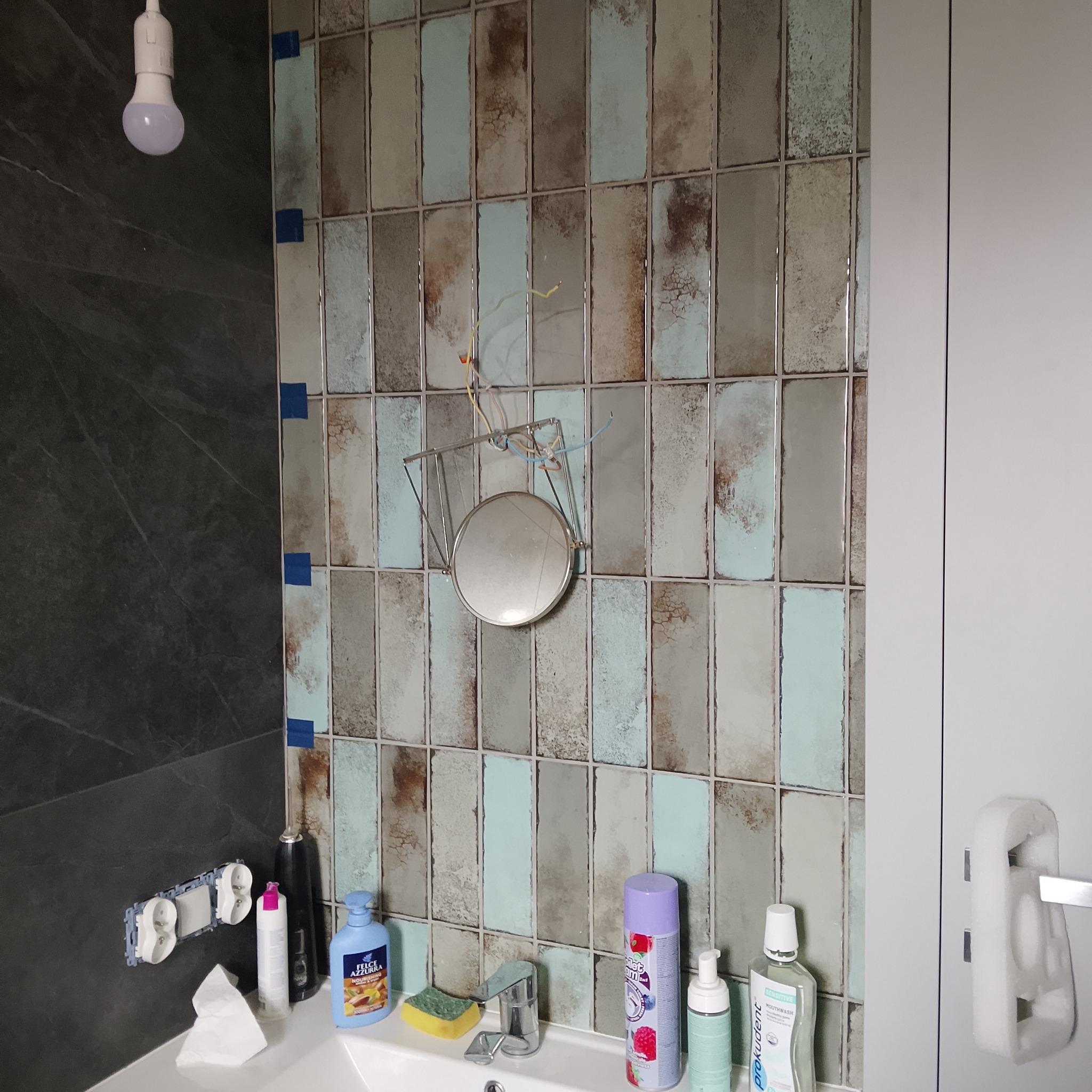 Łazienka w trakcie remontu: położone płytki w odcieniach szarości i błękitu, wiszące lustro bez oświetlenia, widoczne przewody elektryczne.