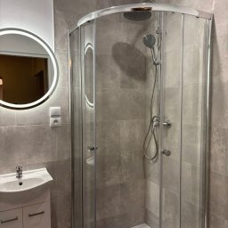 Nowoczesna łazienka z kabiną prysznicową, umywalką z szafką i okrągłym lustrem z podświetleniem LED. Szare płytki na ścianach. Minimalistyczny design.