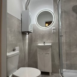 Jasna łazienka z szarymi płytkami, okrągłym lustrem z podświetleniem LED, kabiną prysznicową i podwieszanym piecem gazowym z odprowadzeniem spalin.