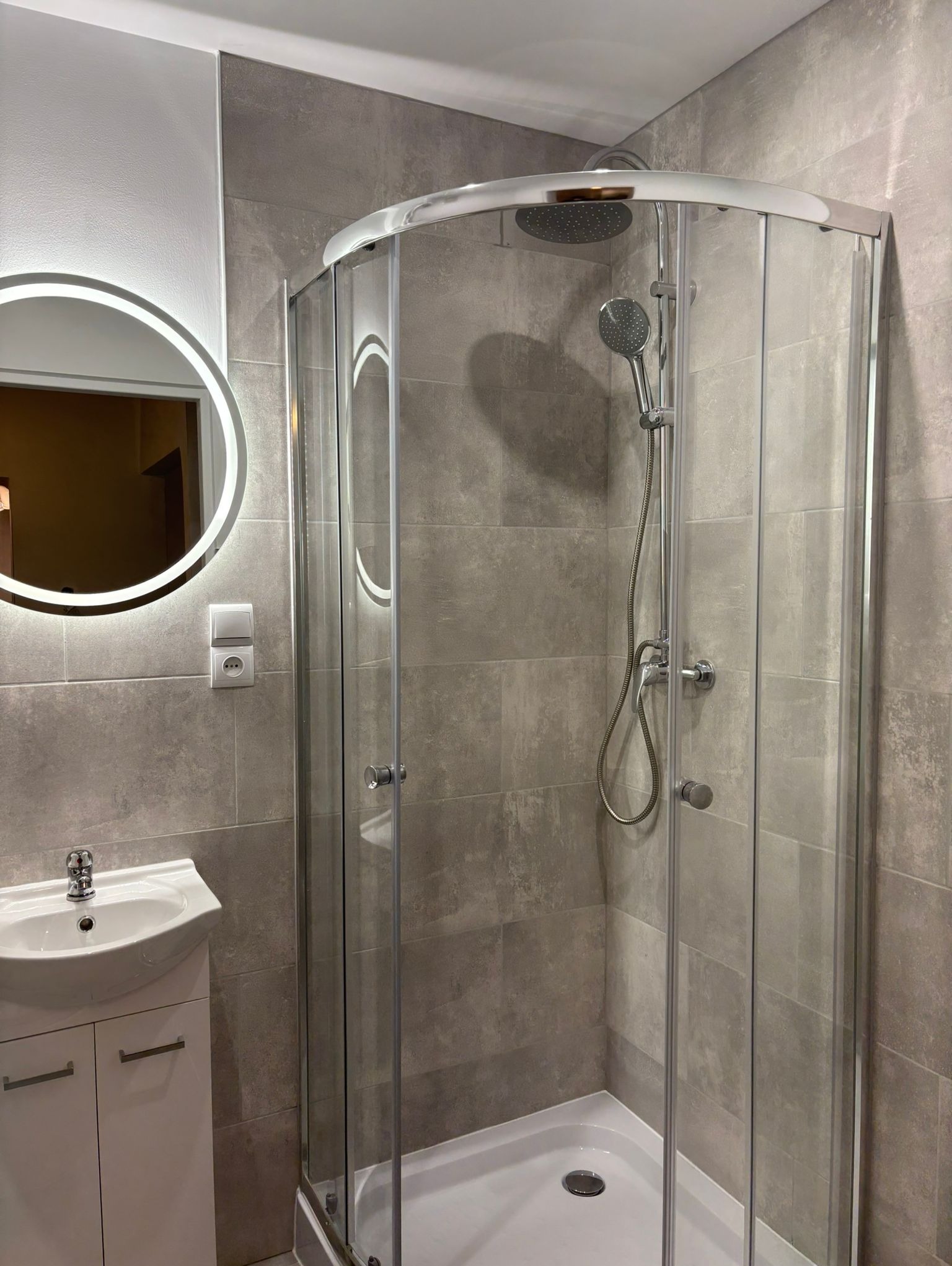 Nowoczesna łazienka z kabiną prysznicową, umywalką z szafką i okrągłym lustrem z podświetleniem LED. Szare płytki na ścianach. Minimalistyczny design.