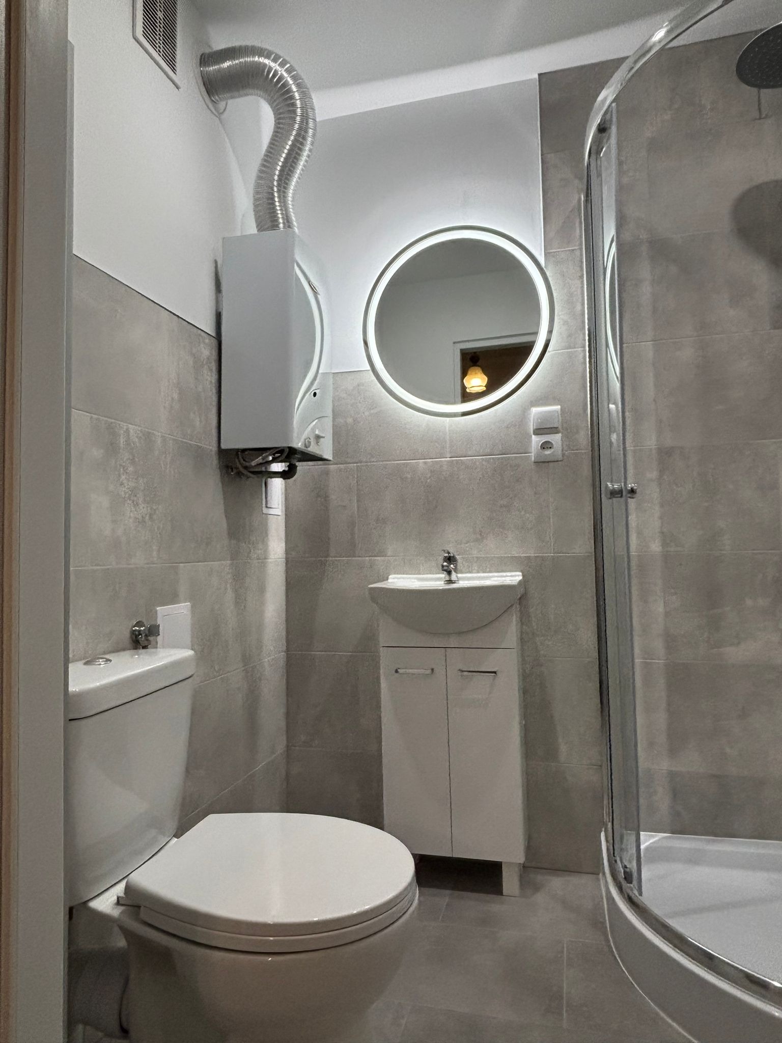 Jasna łazienka z szarymi płytkami, okrągłym lustrem z podświetleniem LED, kabiną prysznicową i podwieszanym piecem gazowym z odprowadzeniem spalin.