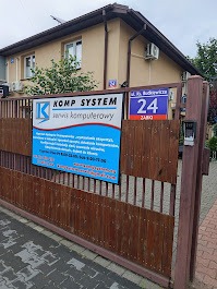 Tablica informacyjna 'Komp System Serwis Komputerowy' na brązowej bramie posesji z adresem 'ul. Budkiewicza 24, Ząbki' na budynku w tle.