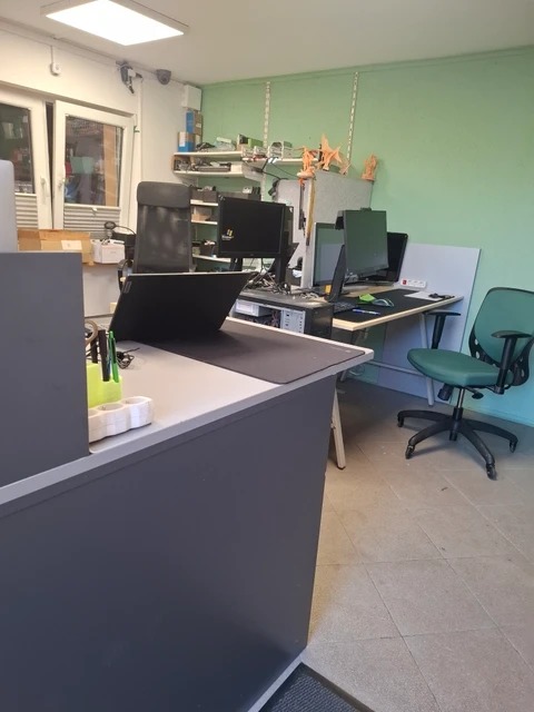 Stanowiska pracy z komputerami w biurze firmy IT. Widoczne monitory, laptopy, biurka i krzesła. Na ścianie półki z akcesoriami komputerowymi.