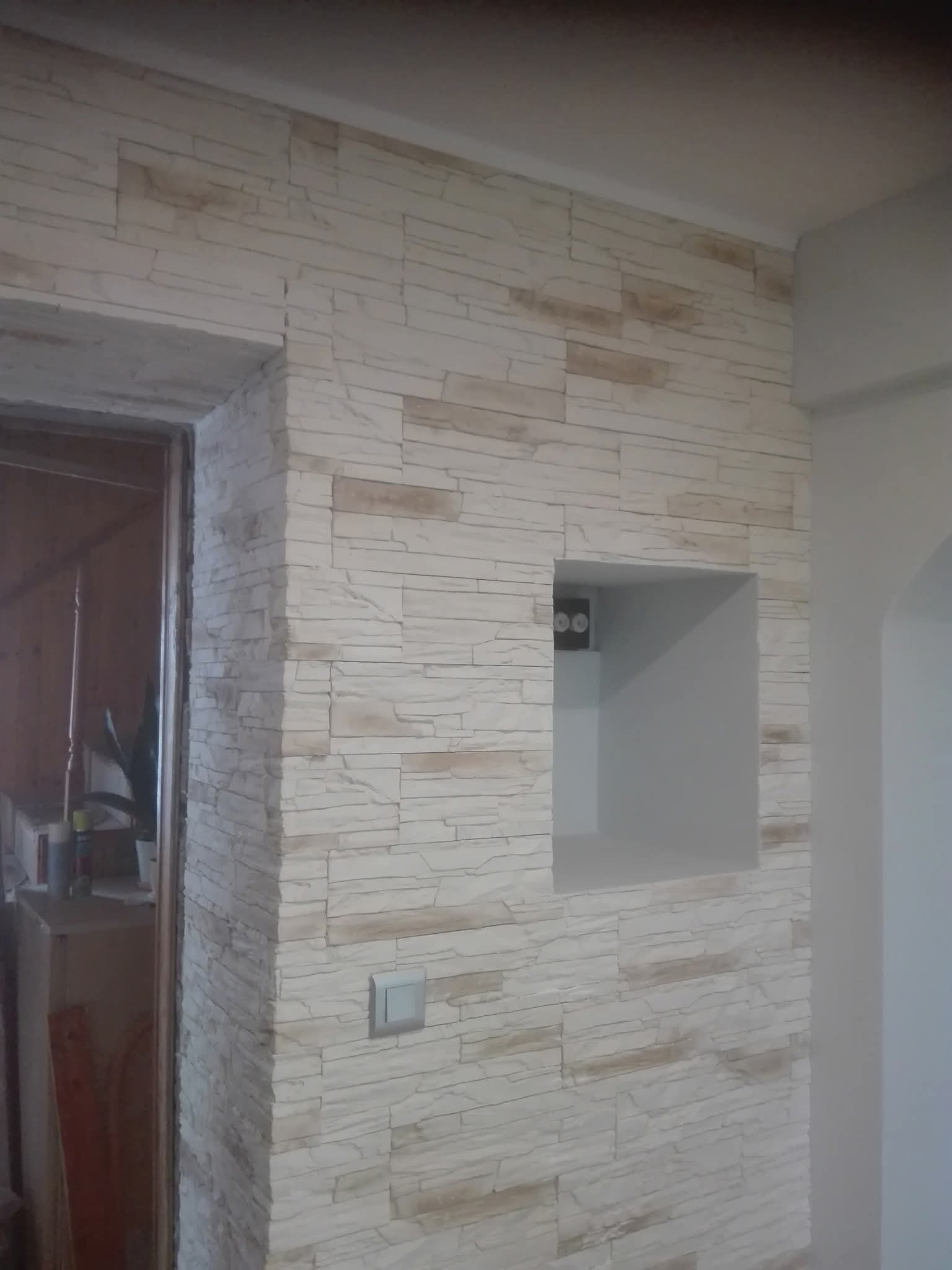 Ściana wykończona dekoracyjną, jasną cegłą z wnęką i włącznikiem światła. Widoczny fragment futryny drzwi. Neutralna kolorystyka, imitacja kamienia.