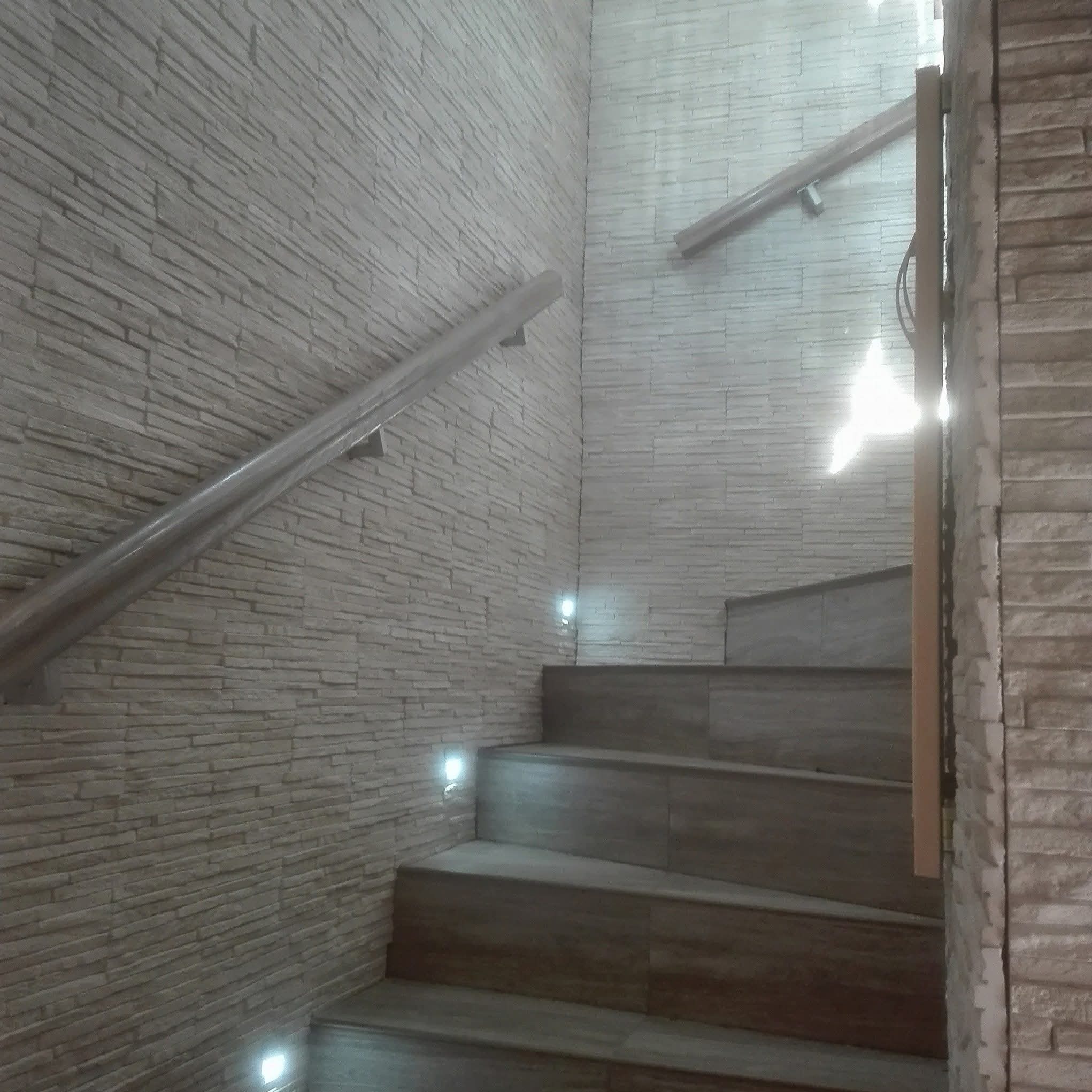 Wykończone schody z kamienną okładziną ścian i metalową poręczą, oświetlone punktowymi lampami. Widoczne detale wykończenia i faktura materiałów.