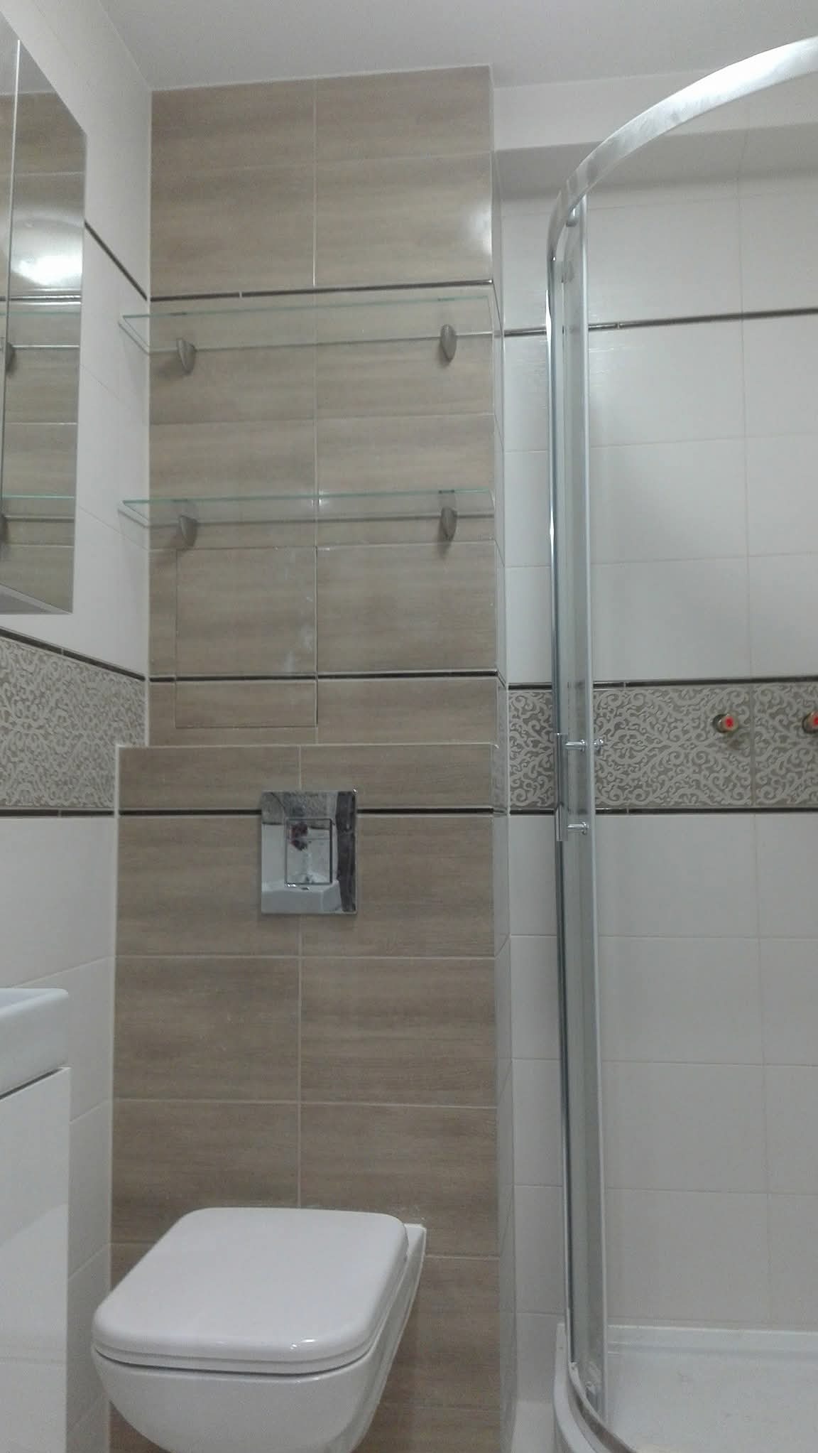 Wykończona łazienka z beżowymi płytkami, szklanymi półkami i kabiną prysznicową. Widoczna podwieszana toaleta i dekoracyjny pas płytek. Nowoczesny design.