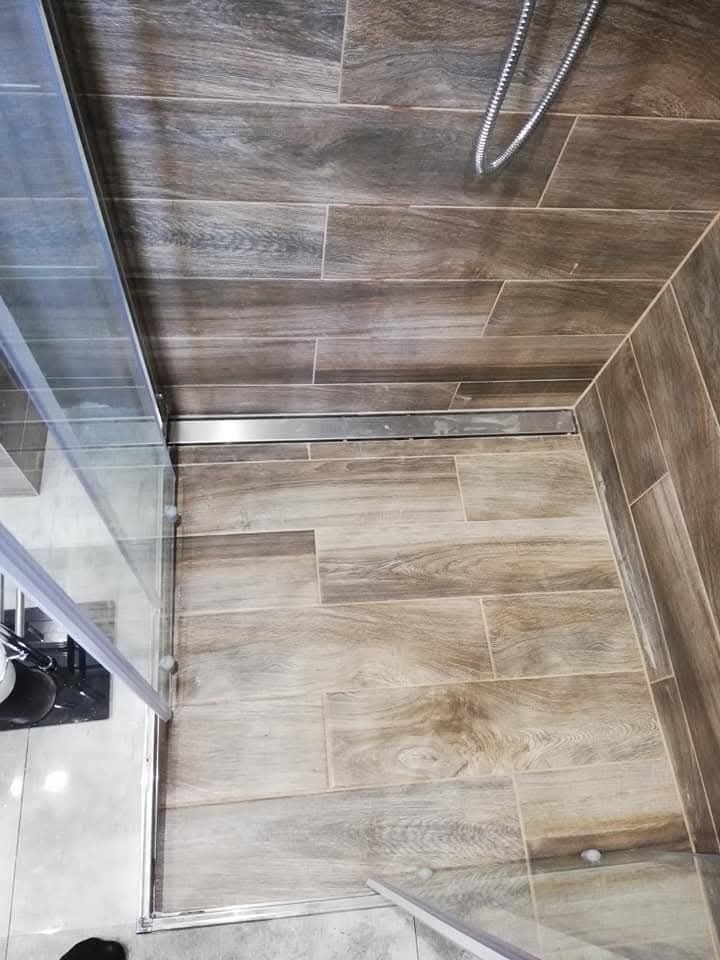 Ukończona kabina prysznicowa z odpływem liniowym i płytkami imitującymi drewno na podłodze i ścianach. Widok z góry, fragment kabiny z szybą.