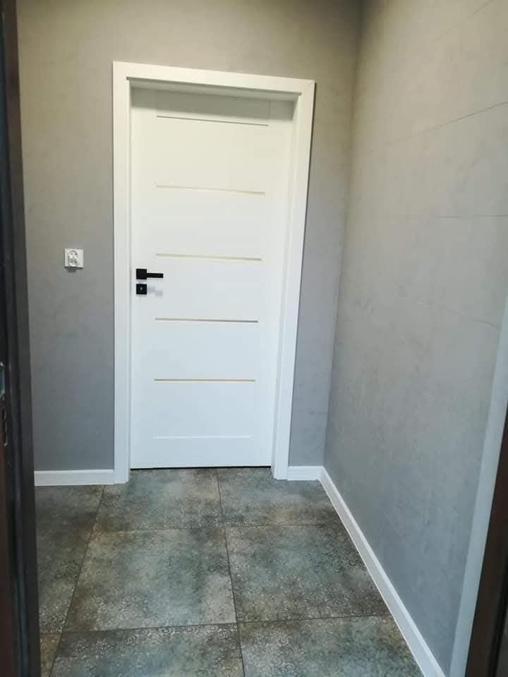 Nowoczesne, białe drzwi z czarną klamką w szarej, wykończonej płytkami łazience. Minimalistyczny design, widoczne detale wykończenia i płytki podłogowe.