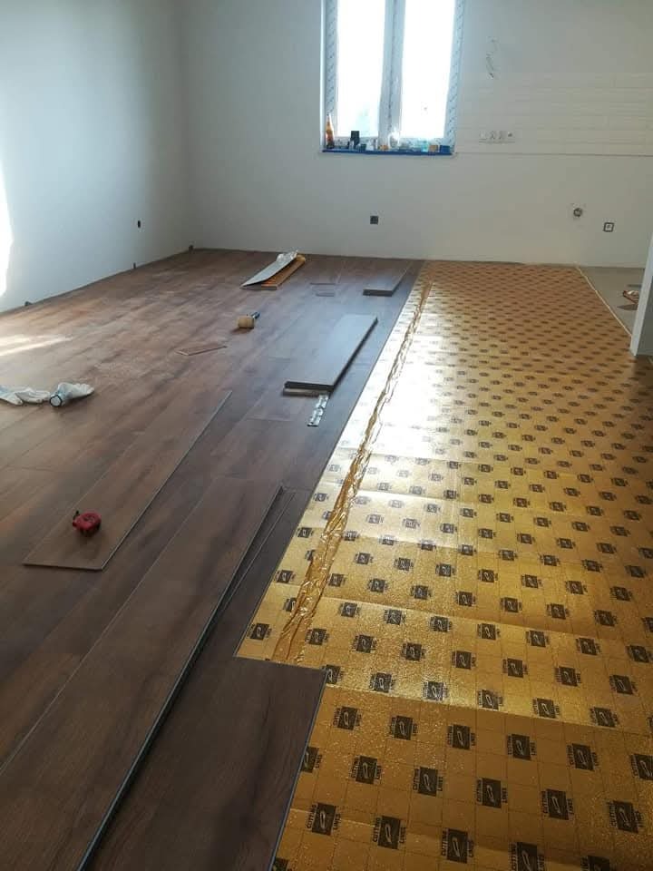 Układanie paneli podłogowych w jasnym pokoju. Częściowo widoczna folia izolacyjna, panele w odcieniach brązu. Narzędzia pracy widoczne w tle.