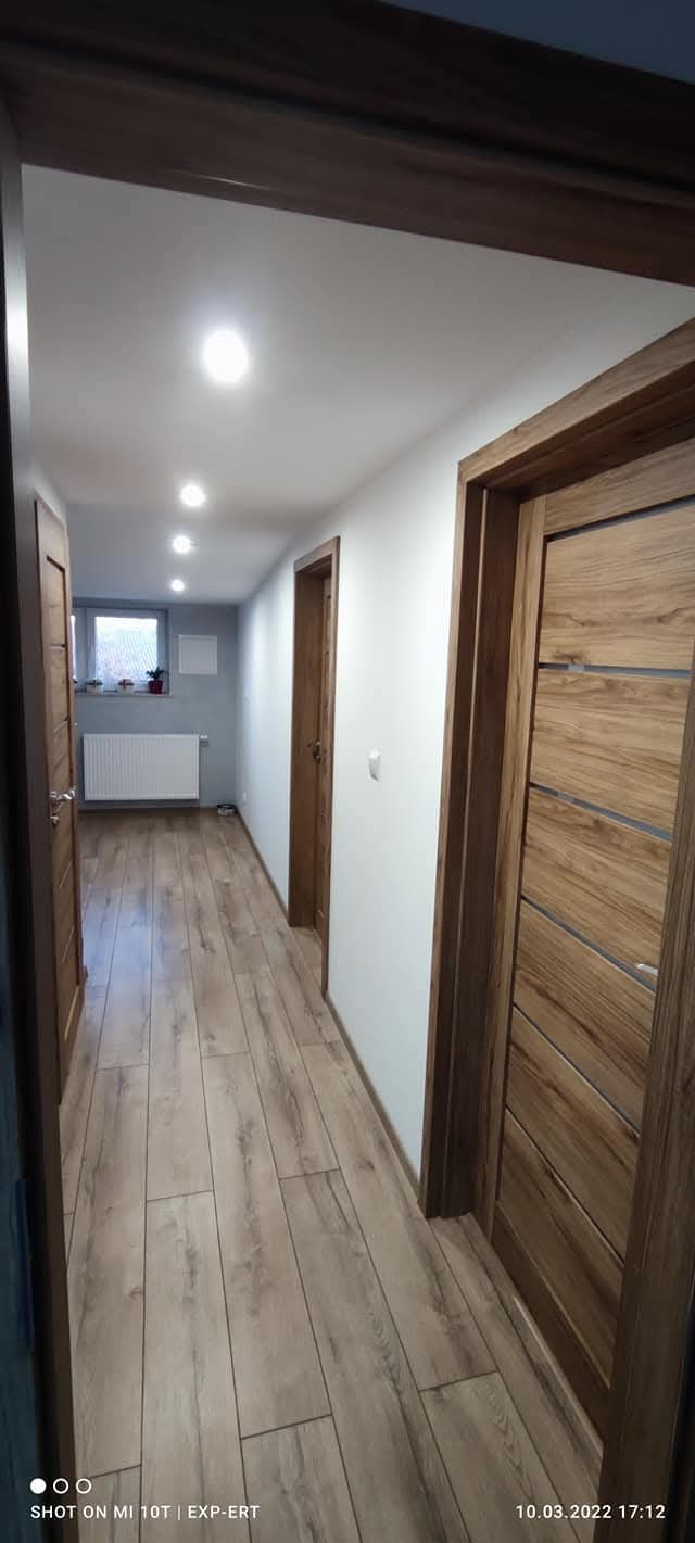 Wykończony korytarz z panelami podłogowymi w odcieniach brązu, białymi ścianami i drewnianymi drzwiami z poziomymi wstawkami. Widoczne oświetlenie sufitowe.