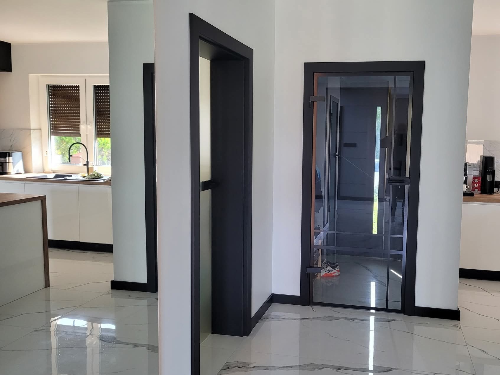 Nowoczesne, czarne drzwi wewnętrzne z przeszkleniami w jasnym wnętrzu. Widoczna marmurowa podłoga i fragment kuchni w tle. Minimalistyczny design.