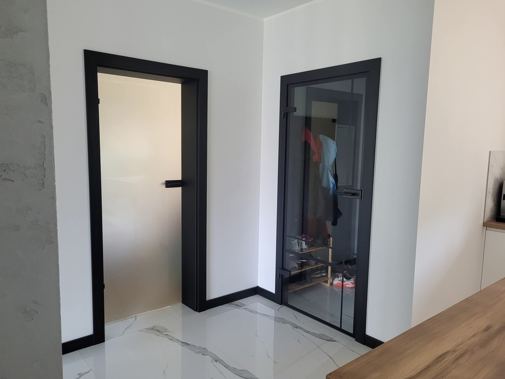 Dwa nowoczesne, czarne obramowania drzwi w jasnym wnętrzu. Jedne drzwi szklane, drugie pełne. Marmurowe płytki podłogowe z szarymi żyłami.