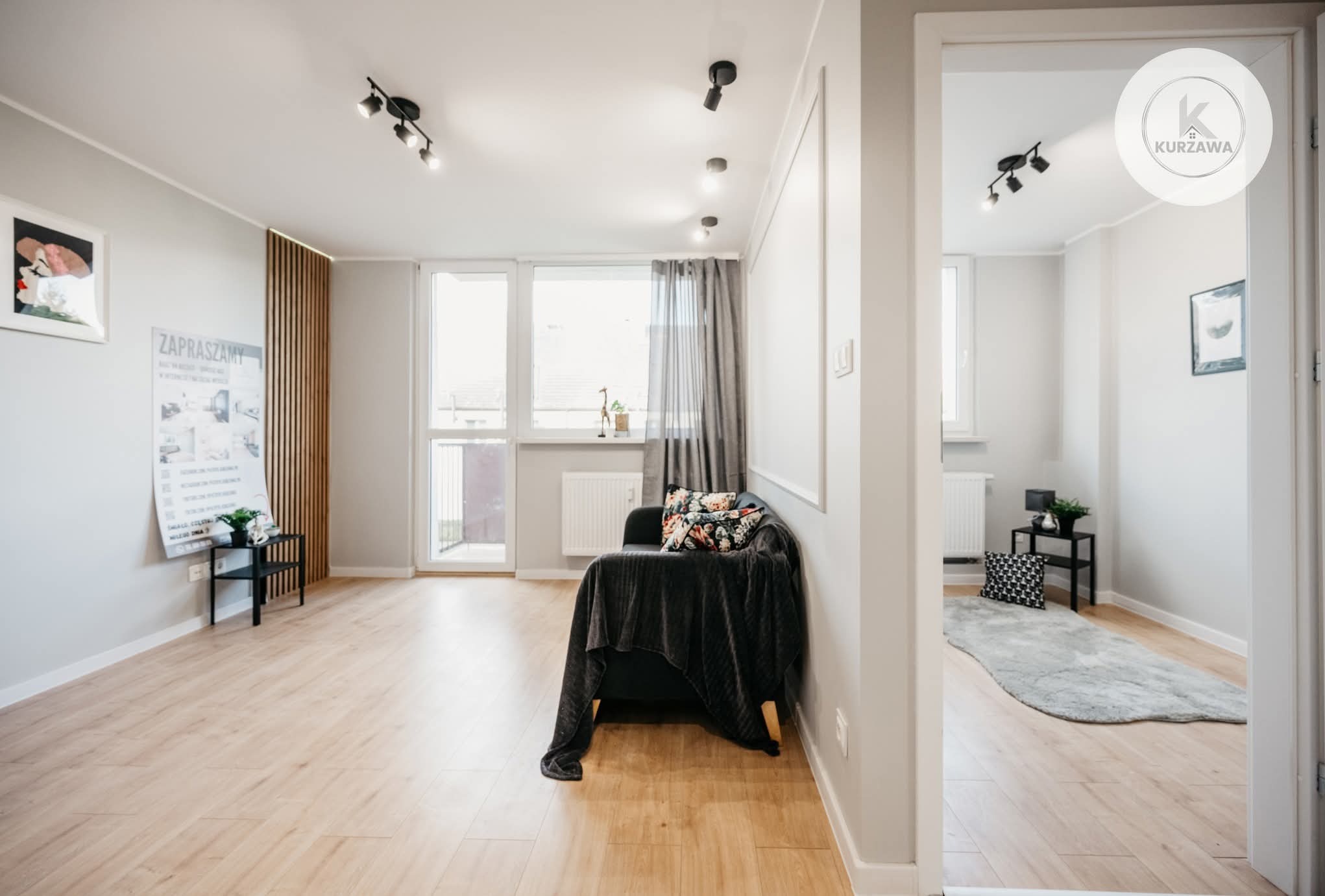 Jasne wnętrze salonu po remoncie: drewniana podłoga, szara sofa z narzutą, okno balkonowe z zasłonami, oświetlenie sufitowe. Widok na drugie pomieszczenie.