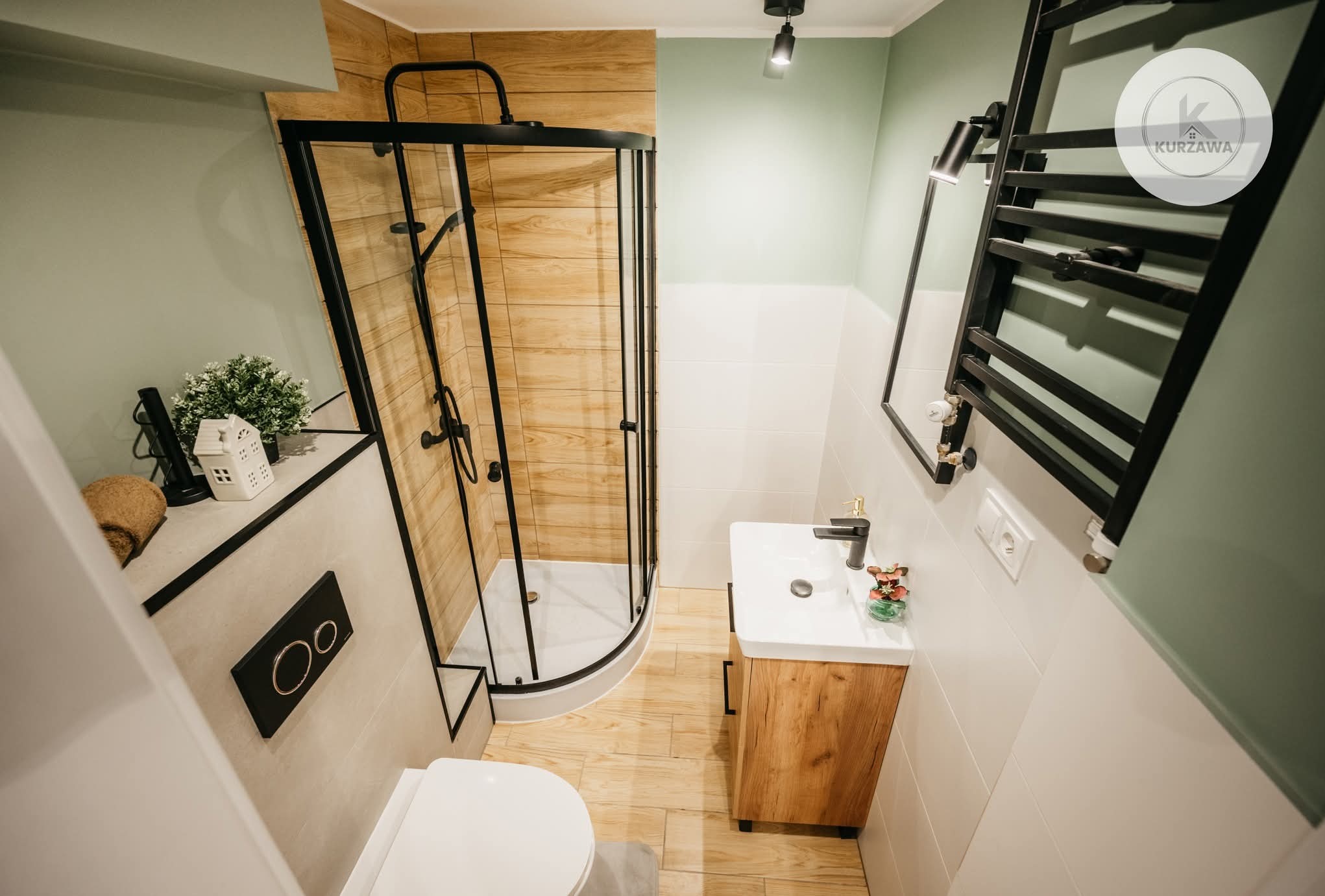 Nowoczesna łazienka po remoncie w Warszawie: kabina prysznicowa z czarnymi profilami, drewnopodobne płytki, umywalka z szafką i grzejnik drabinkowy.