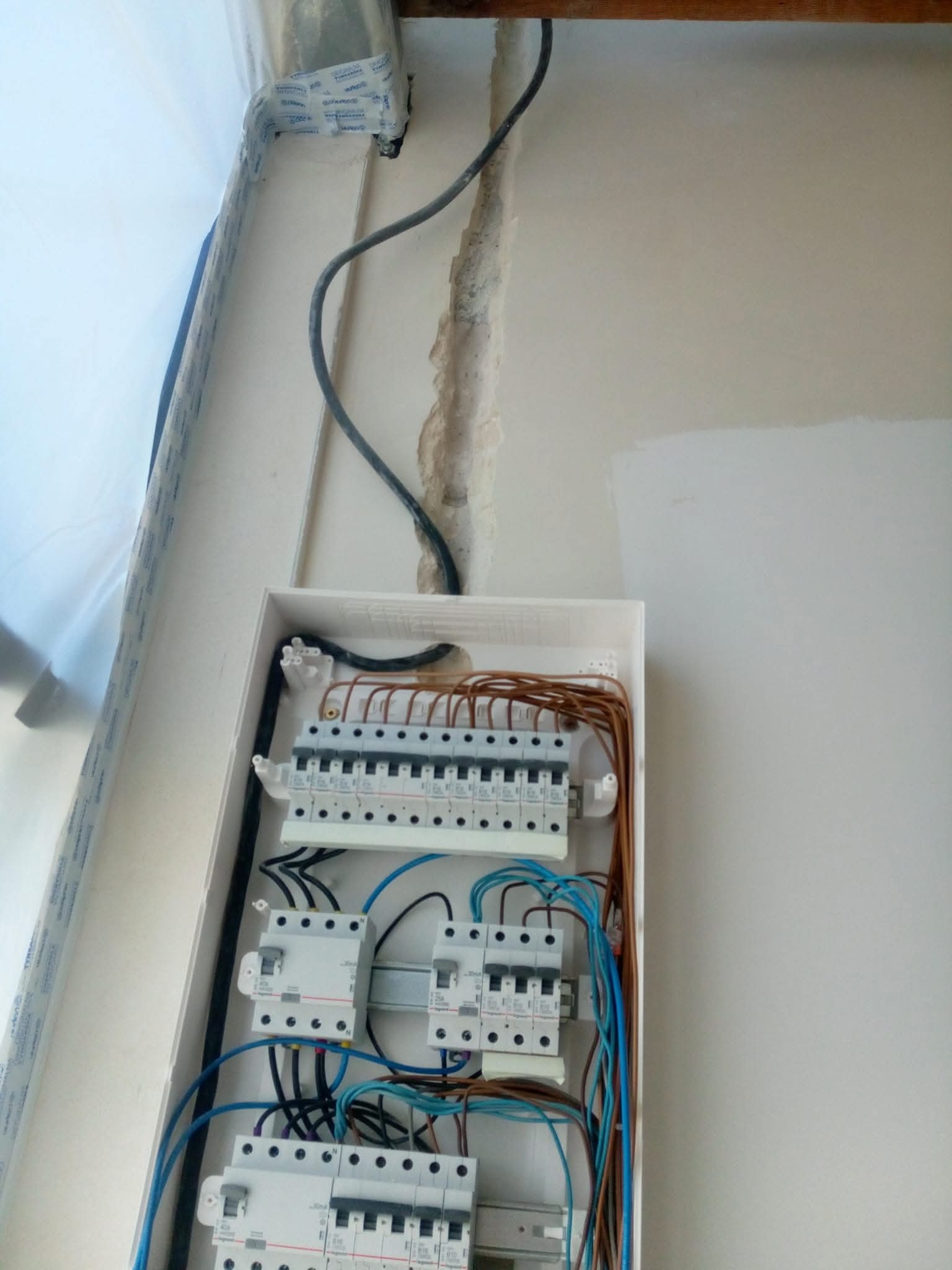 Rozdzielnica elektryczna w białej obudowie, widoczne kable i bezpieczniki. Ściana w trakcie malowania, widoczne niedomalowane fragmenty. Kabel wystaje ze ściany.