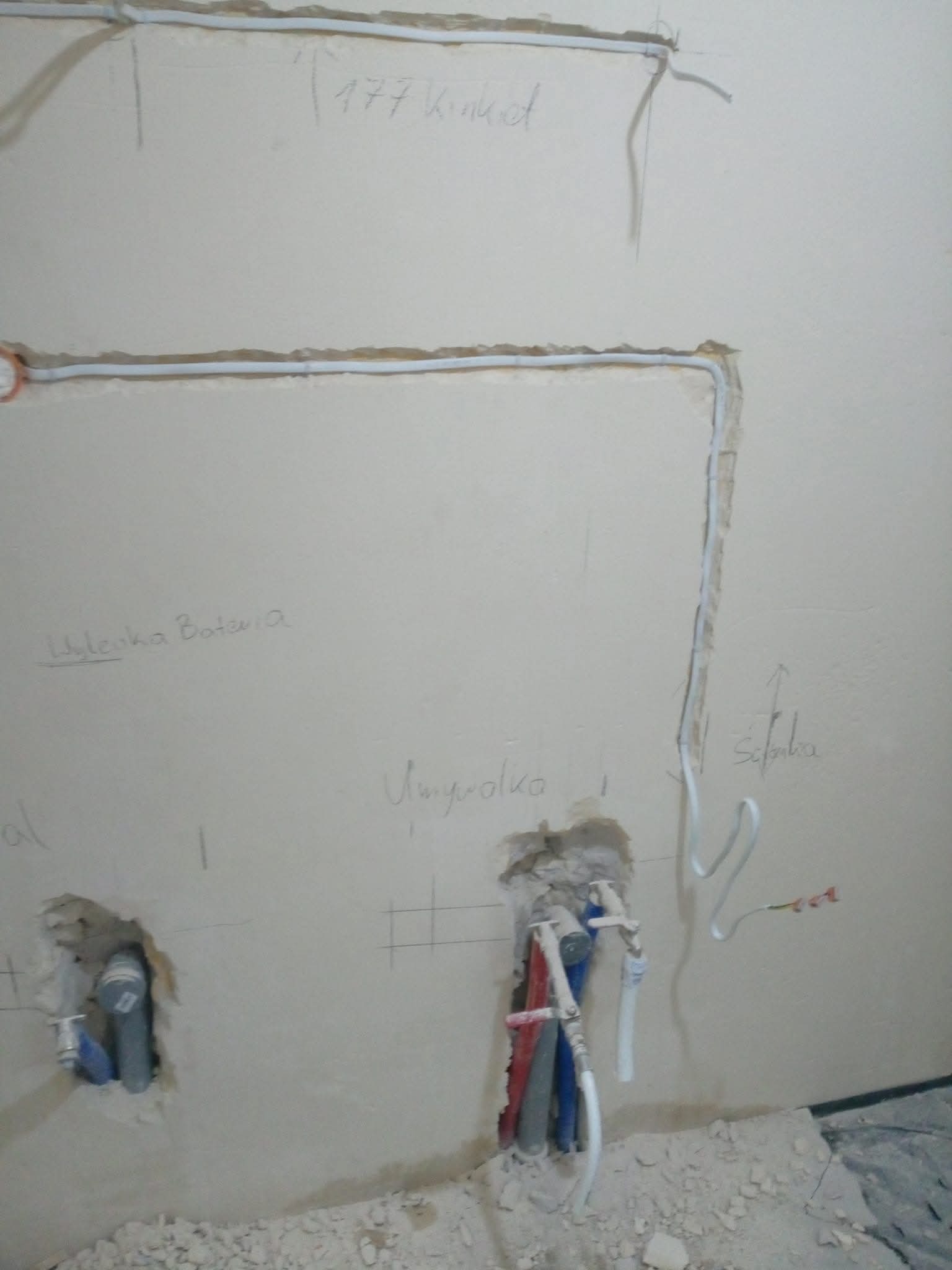 Ściana z instalacją elektryczną i wodną w trakcie montażu. Widoczne kable i rury, oznaczenia na ścianie. Prace instalacyjne w surowym stanie.