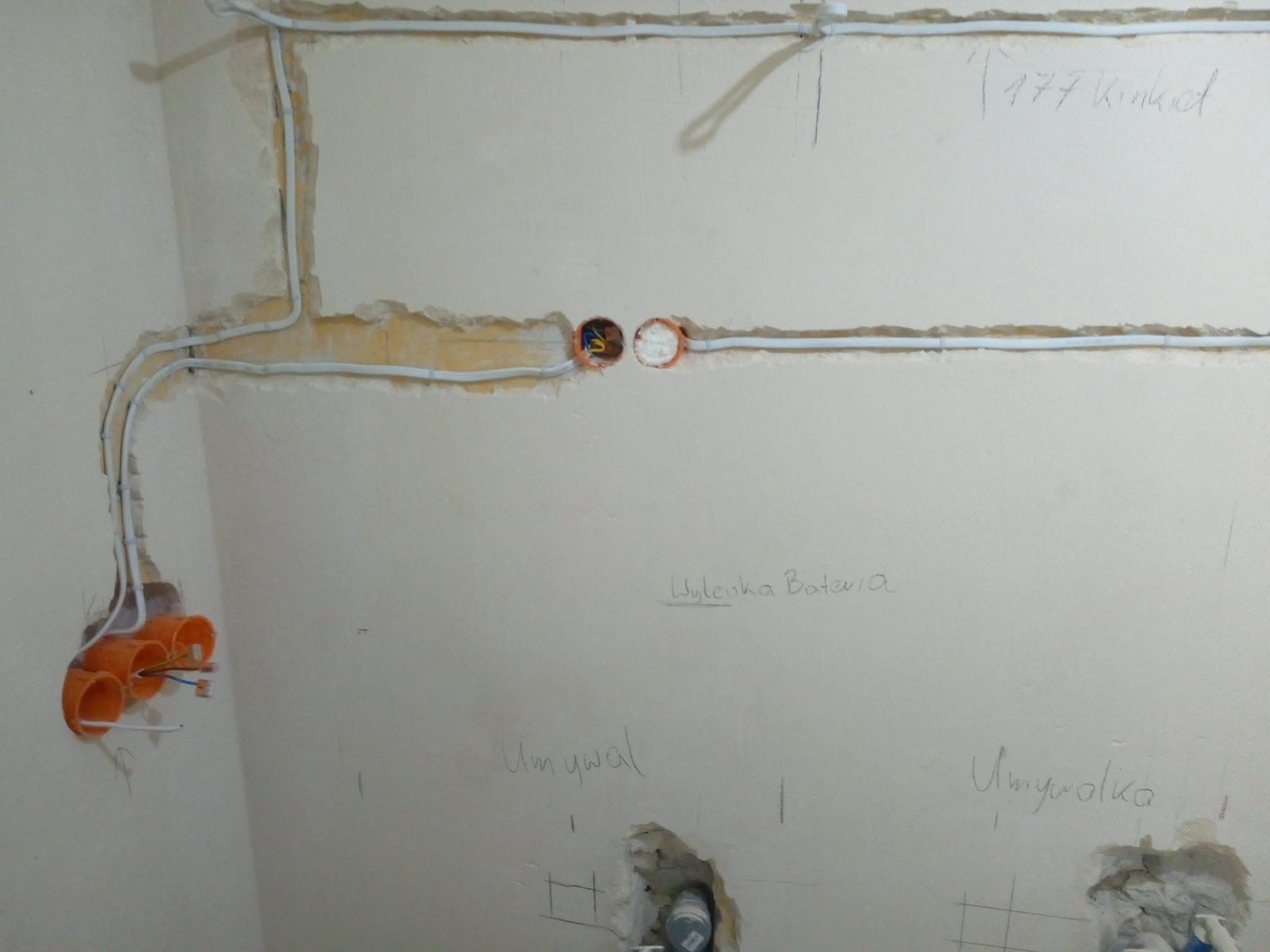 Ściana w trakcie prac elektrycznych: widoczne kable w peszlach, puszki instalacyjne i notatki ołówkiem na ścianie. Etap prac instalacyjnych wewnątrz budynku.