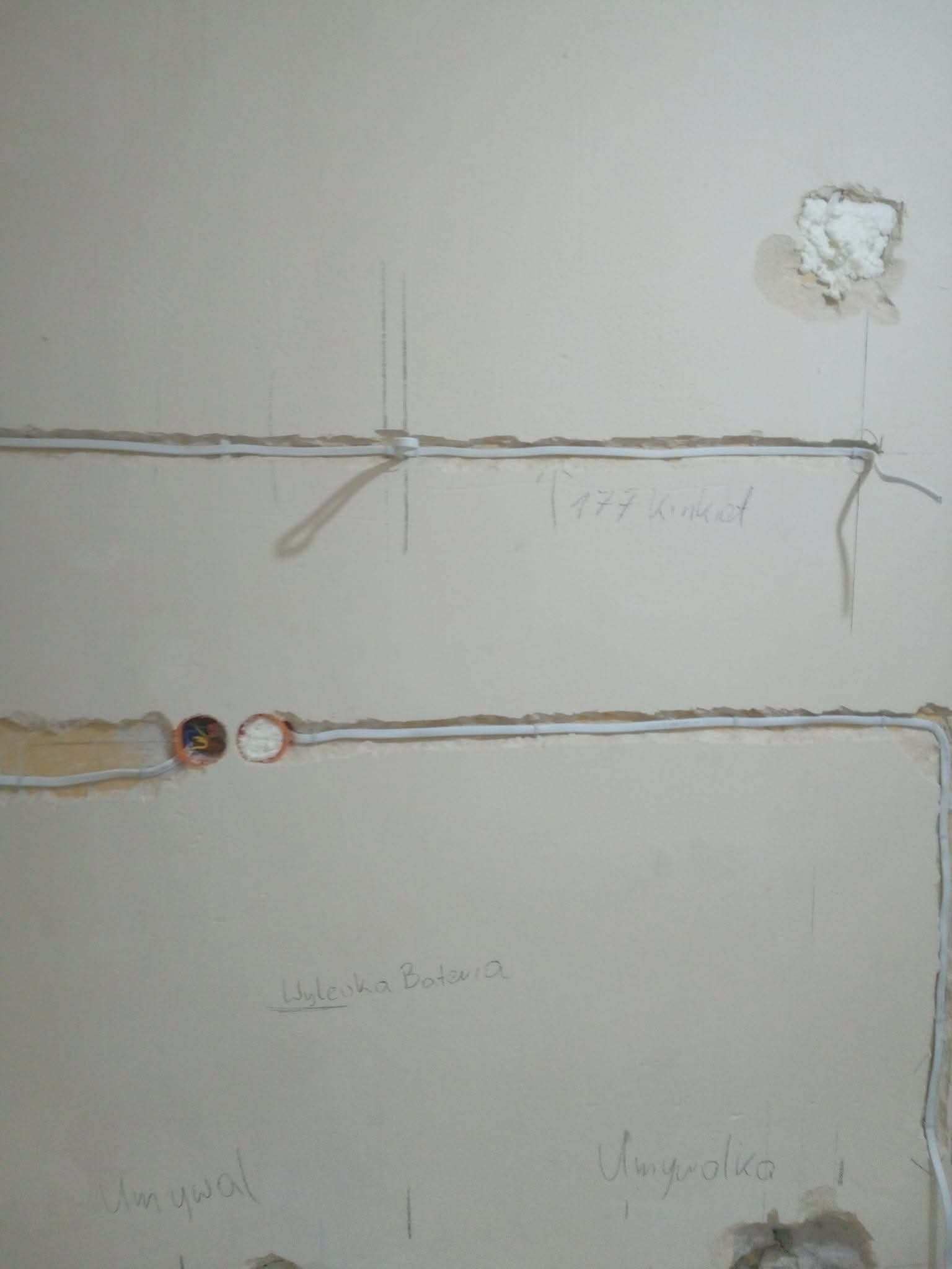 Ściana z widocznymi przewodami elektrycznymi w bruzdach, przygotowana do dalszych prac wykończeniowych. Widoczne napisy ołówkiem na ścianie.