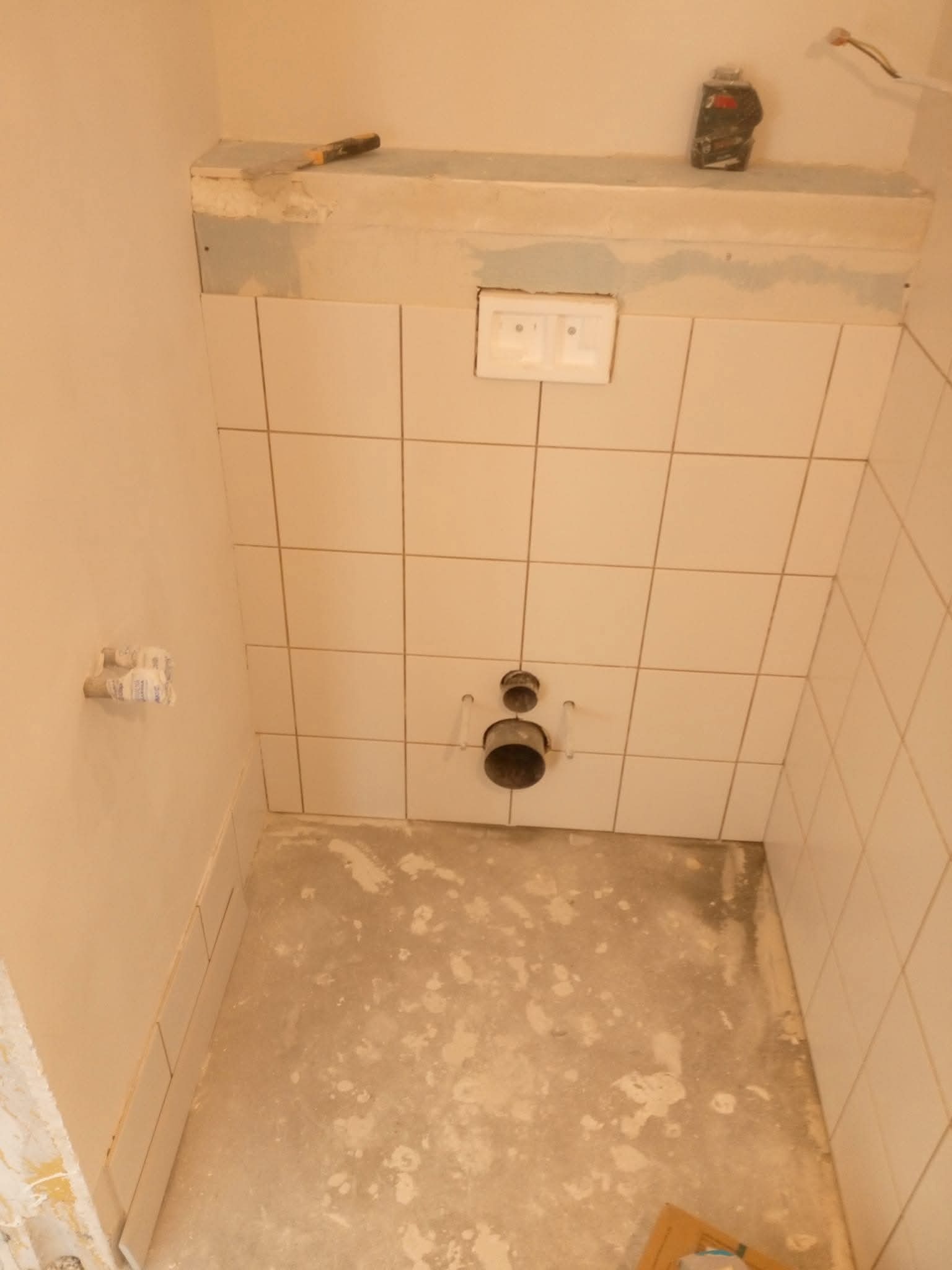 Wnętrze łazienki w trakcie remontu: ułożone jasne płytki na ścianie, widoczne rury odpływowe, surowa podłoga. Na górze narzędzie i akcesoria glazurnicze.