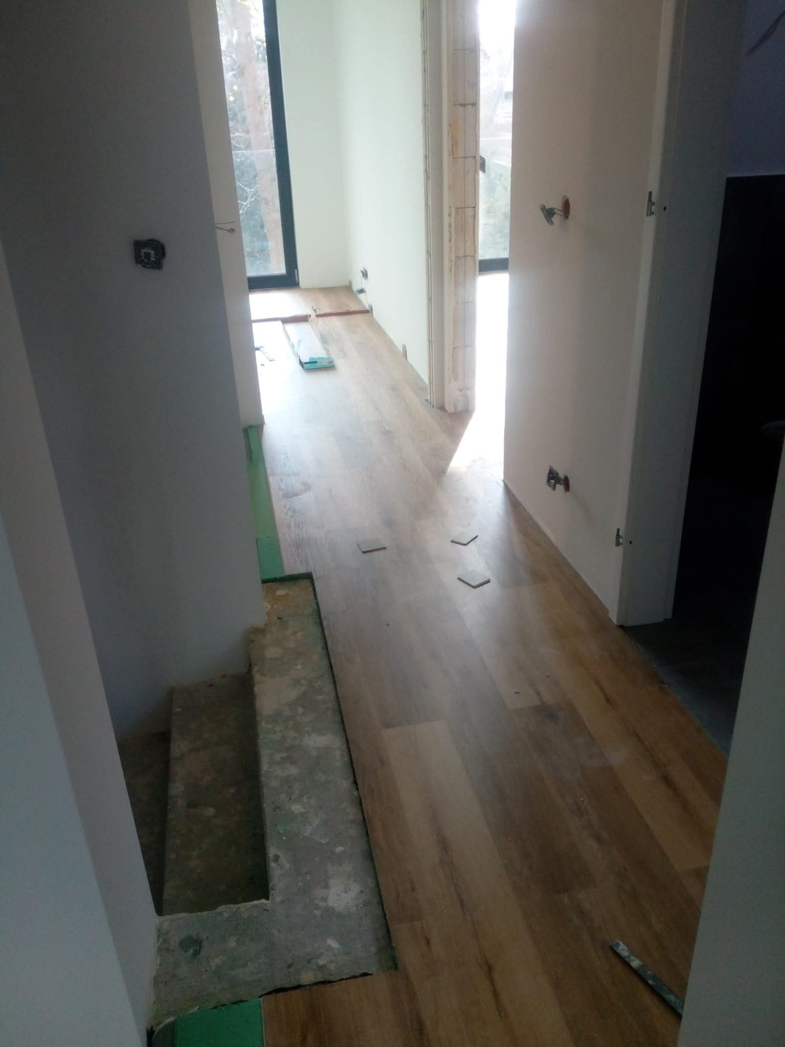 Układanie paneli podłogowych w jasnym wnętrzu. Widoczne fragmenty podkładu i niedokończone prace przy schodach. Naturalne światło wpada przez okno.