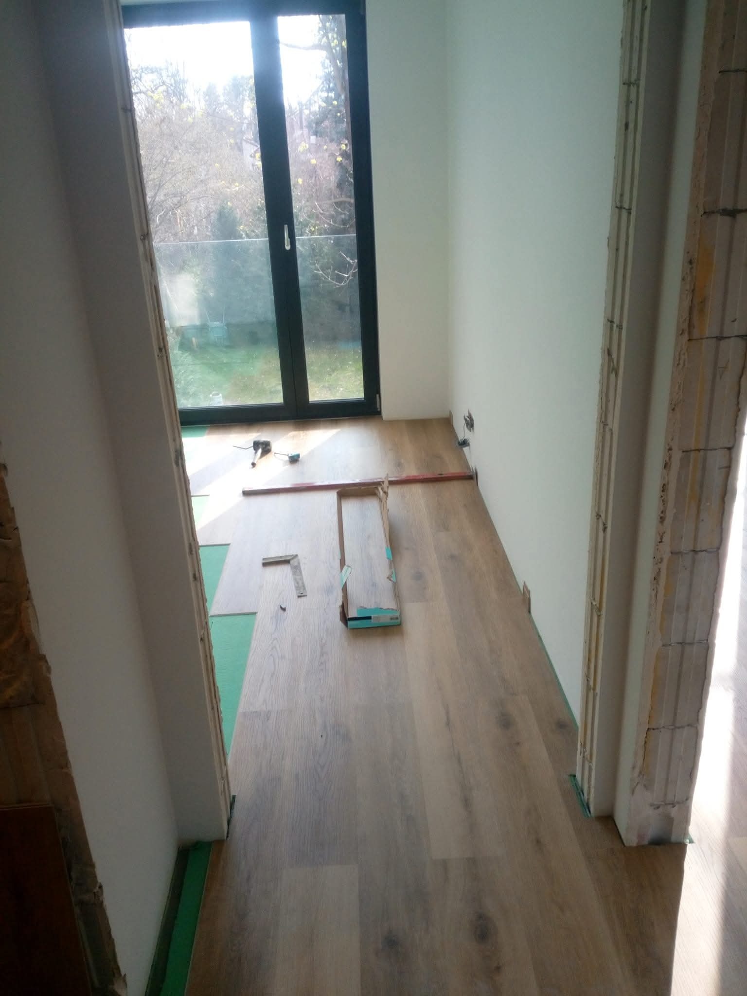 Układanie paneli podłogowych w jasnym wnętrzu z oknem balkonowym. Widoczne narzędzia i fragmenty paneli. Podkład pod panele w kolorze zielonym.