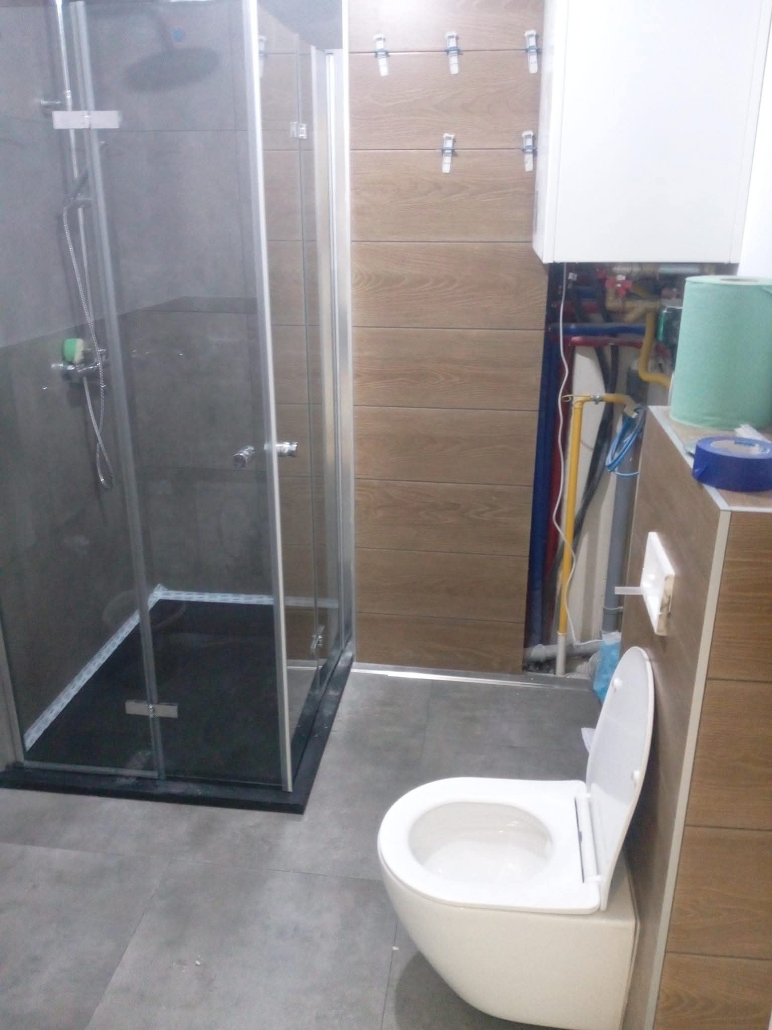 Nowoczesna łazienka z kabiną prysznicową typu walk-in, podwieszaną toaletą i płytkami imitującymi drewno na ścianie. Widoczne instalacje za zabudową.