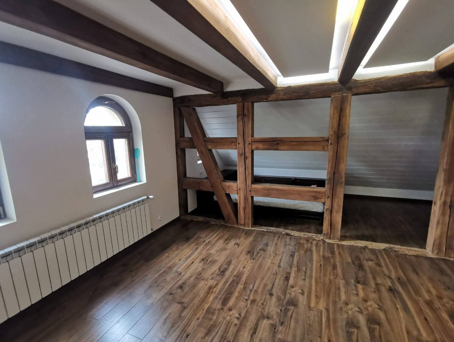 Jasne wnętrze z drewnianymi belkami stropowymi i podłogą. Okno łukowe z grzejnikiem. Konstrukcja z belek dzieli przestrzeń. Oświetlenie LED w suficie.