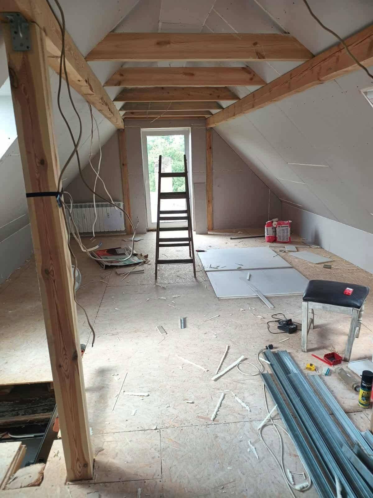 Poddasze w trakcie adaptacji: widoczne drewniane belki, płyty gipsowe, drabina, narzędzia i materiały budowlane. Prace wykończeniowe w surowym wnętrzu.